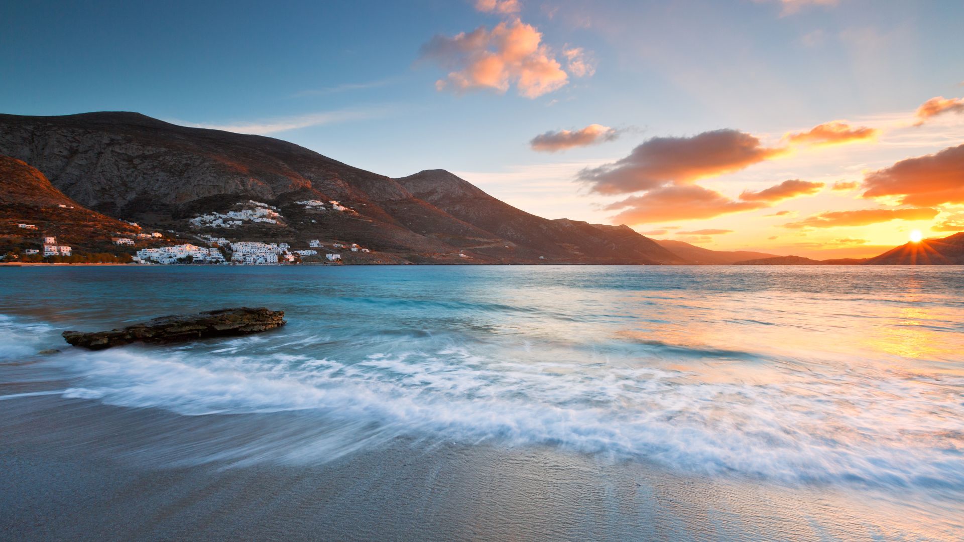 Sunset view of a beach in Amorgos, Greece, with mountains and a village in the background.