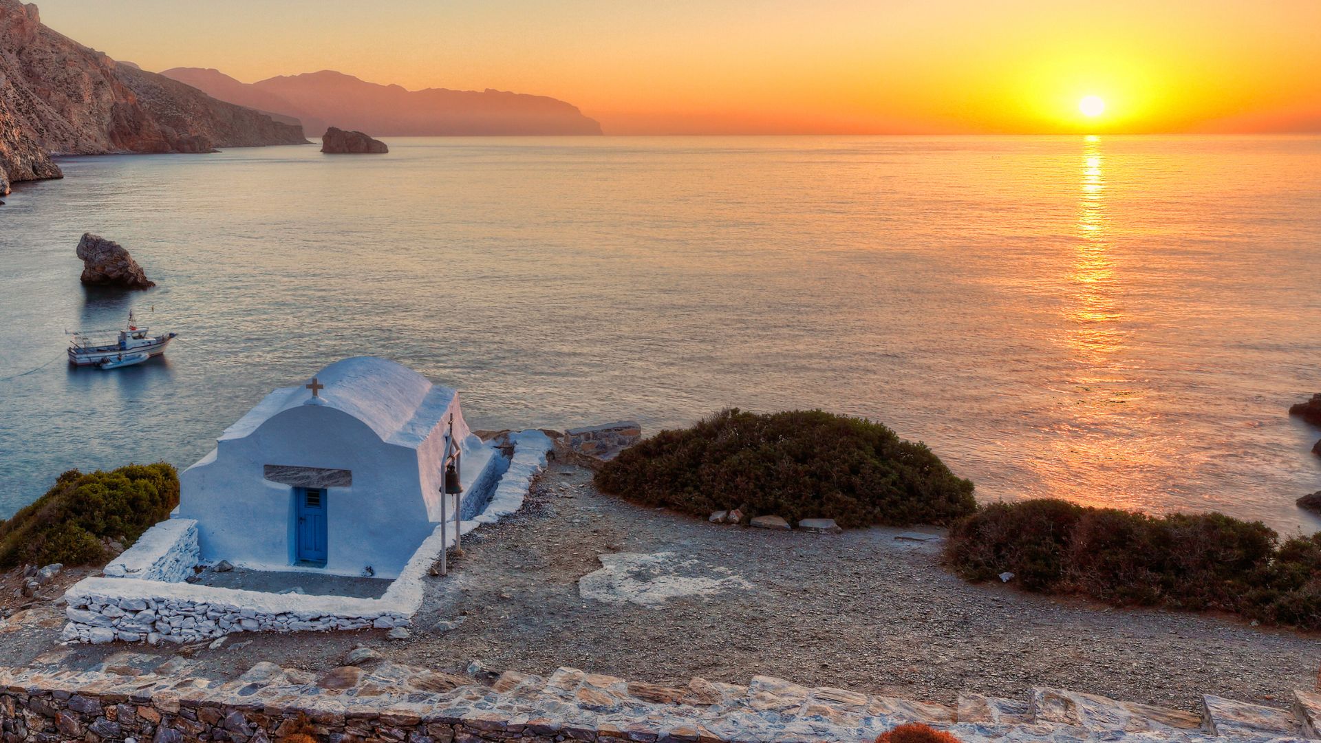 A small white chapel sits on a rocky beach overlooking the Aegean Sea at sunset.