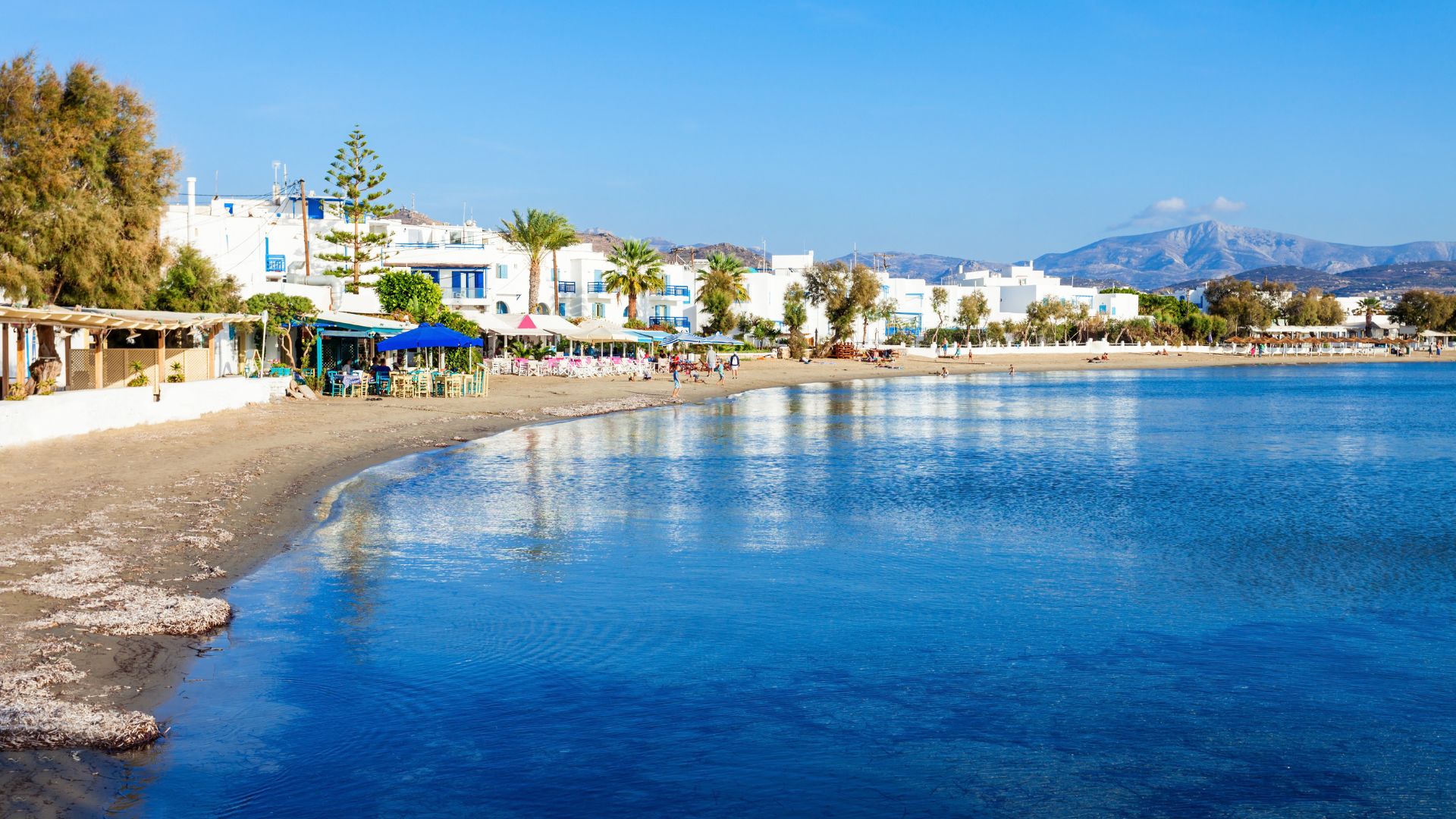 A wide shot of Agios Georgios Beach in Naxos, showing the clear blue water, sandy beach, and white buildings along the shore.