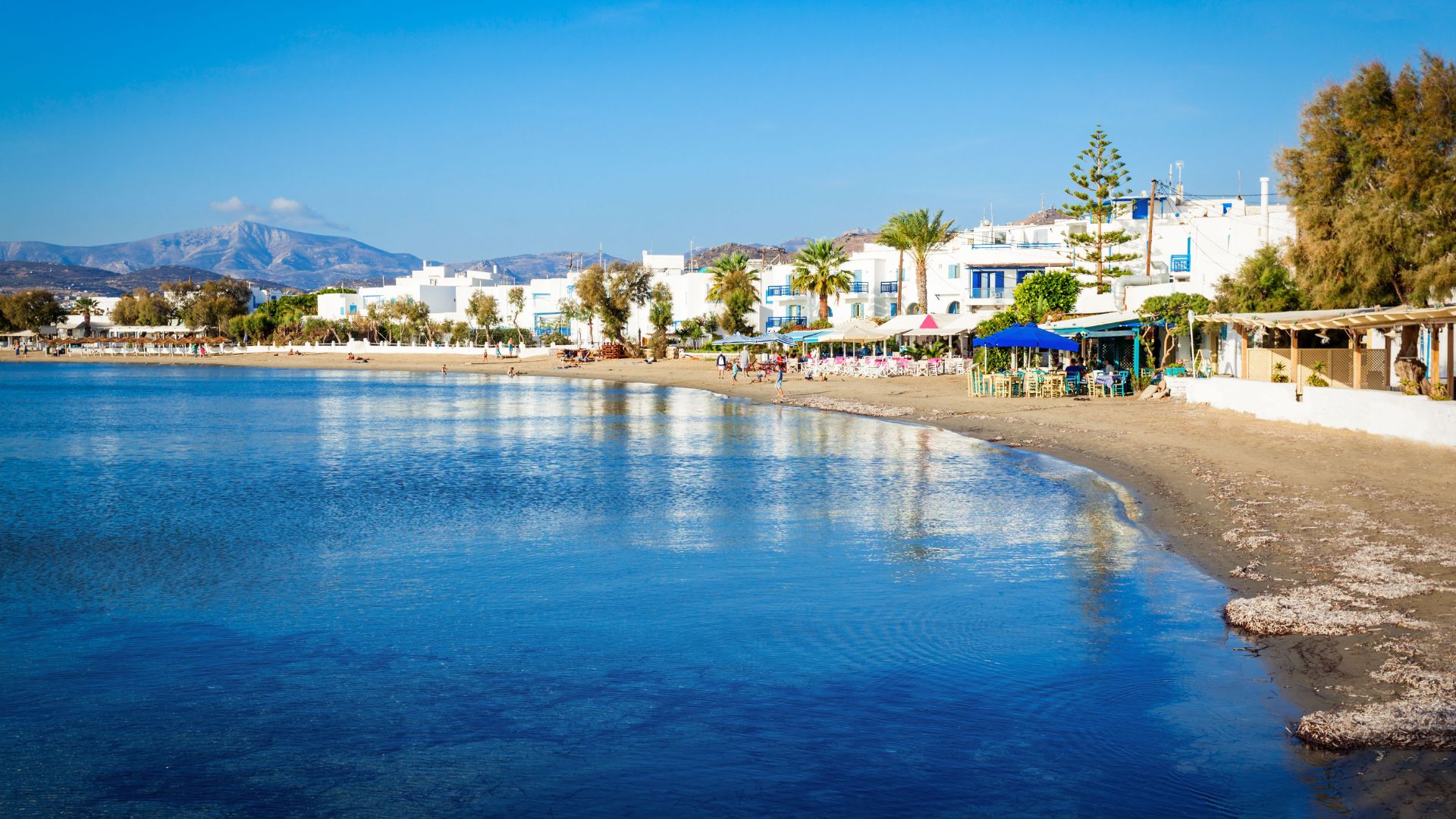 A wide shot of Agios Georgios Beach in Naxos, showing the clear blue water, sandy beach, and white buildings along the shore.