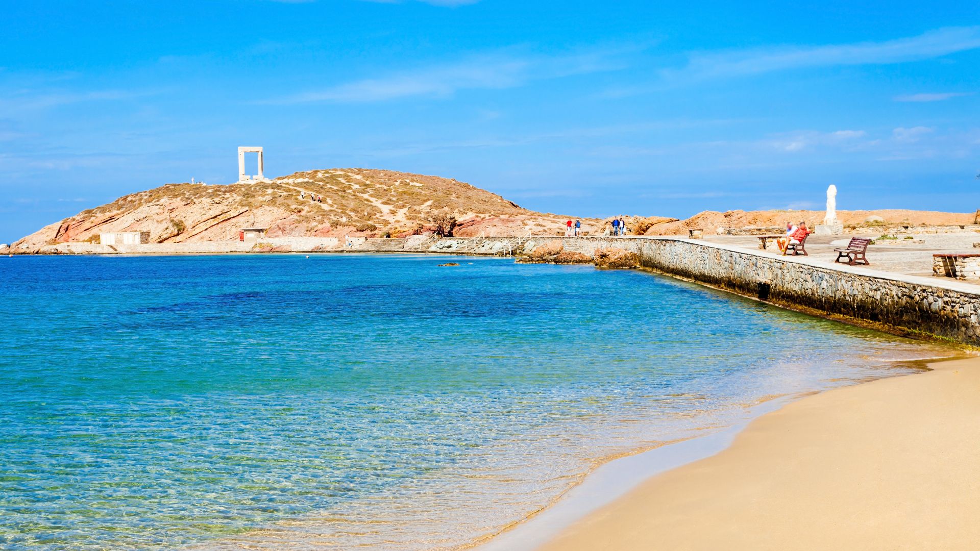 A wide shot of Agios Georgios Beach in Naxos, showing the clear blue water, sandy beach.