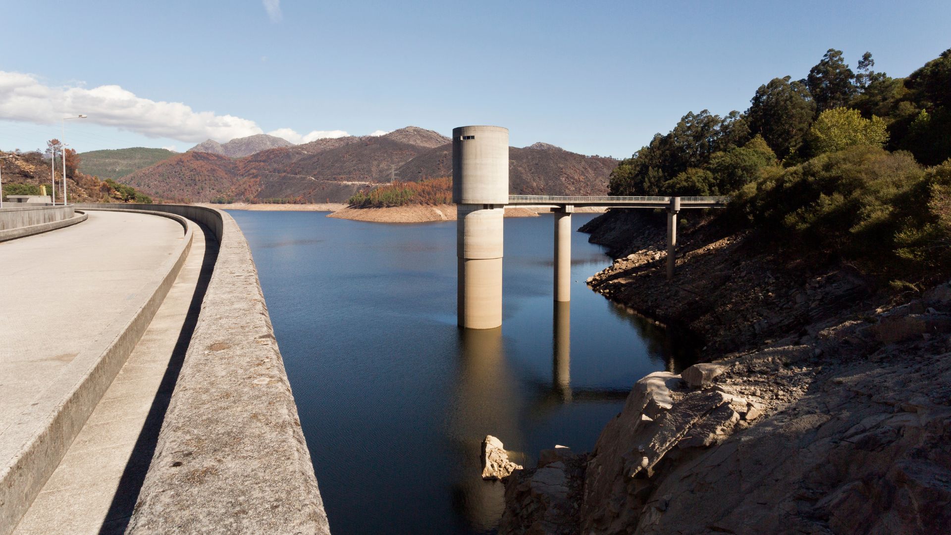 A concrete dam and its water intake tower stand in a large reservoir surrounded by mountains and trees under a clear sky.