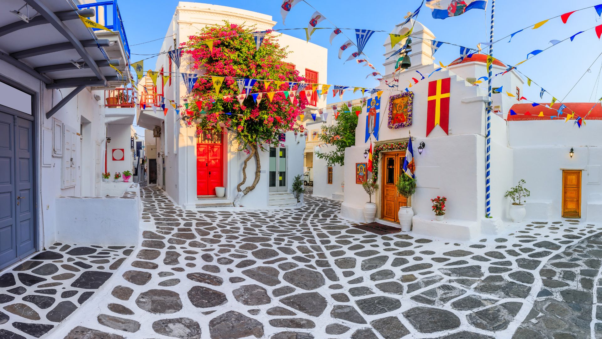 A charming street scene in Ano Mera, Mykonos, featuring white buildings, a stone-paved road, and colorful decorations.