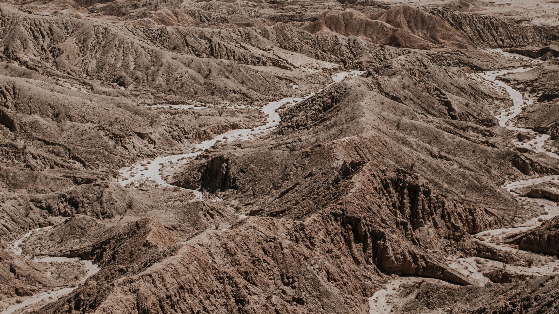 An aerial view of a vast, arid desert landscape featuring numerous eroded, brown hills and a winding dry wash or riverbed carving through the terrain. The scene depicts the characteristic badlands and intricate geological formations of a desert environment.