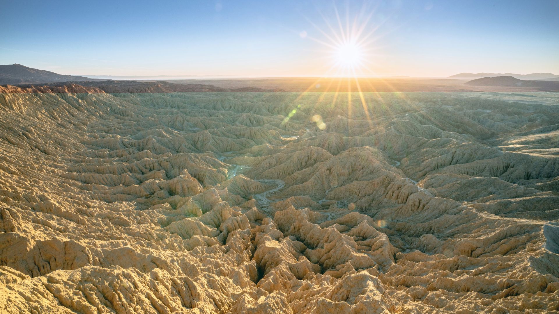 A panoramic view of the Anza-Borrego Desert's badlands at sunrise or sunset, featuring intricate, eroded formations stretching towards the horizon under a clear sky with a brilliant sunburst.