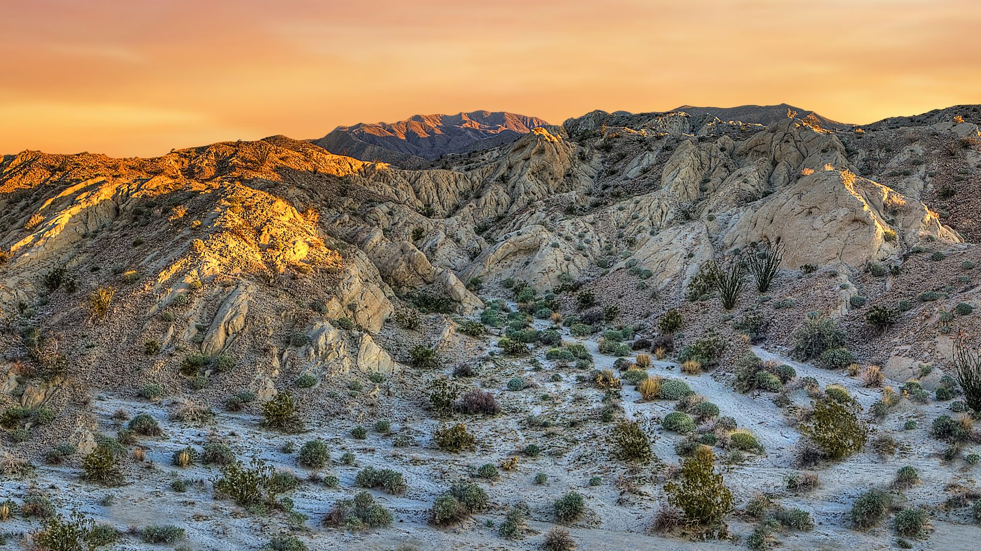 A scenic view of Anza-Borrego Desert State Park in Southern California, showcasing its rugged terrain, mountains, and desert vegetation under an orange sky at sunset.