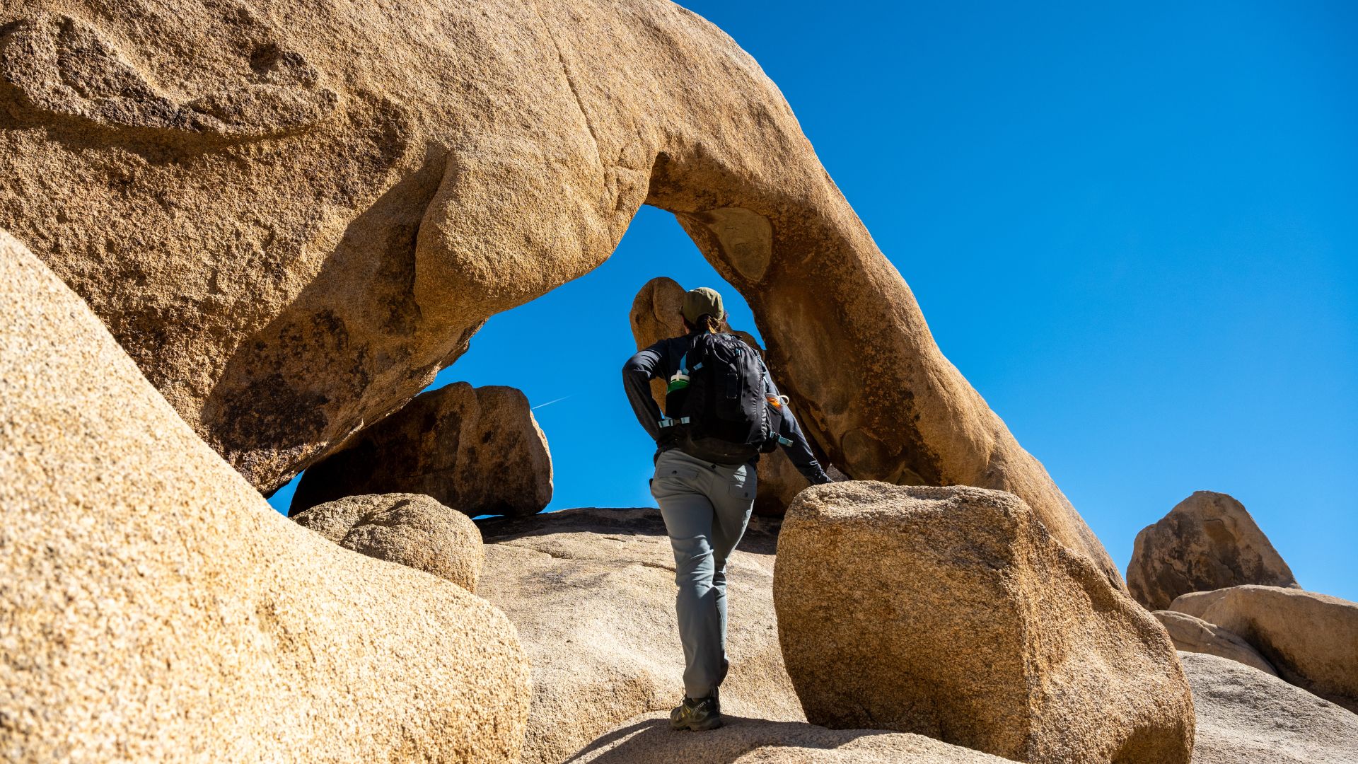 A natural rock arch formation, known as Arch Rock, stands prominently against a clear blue sky in Joshua Tree National Park, California. The arch is composed of large, weathered granite boulders, forming a distinctive curved opening. Surrounding the arch are other large rock formations and sparse desert vegetation under a bright, sunny sky.