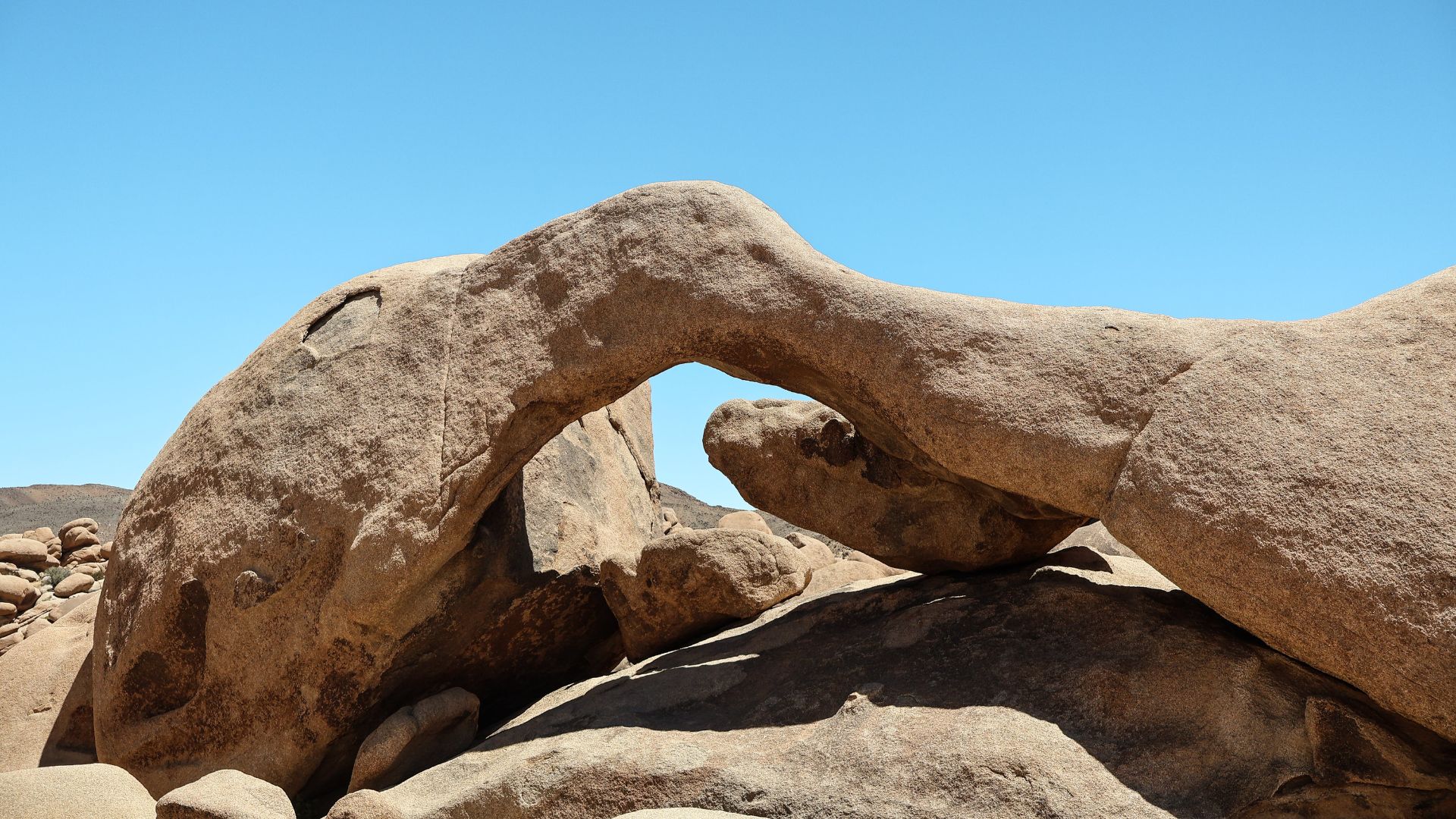A natural rock arch formation, known as Arch Rock, stands prominently against a clear blue sky in Joshua Tree National Park, California. The arch is composed of large, weathered granite boulders, forming a distinctive curved opening. Surrounding the arch are other large rock formations and sparse desert vegetation under a bright, sunny sky.