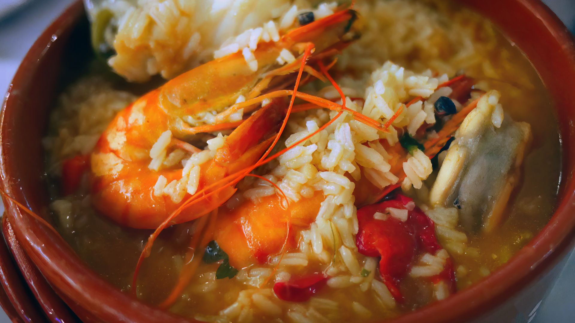A close-up of Arroz de Marisco, a Portuguese seafood rice dish, served in a terracotta bowl. The dish features plump shrimp, rice, and other seafood in a savory broth, garnished with red peppers.