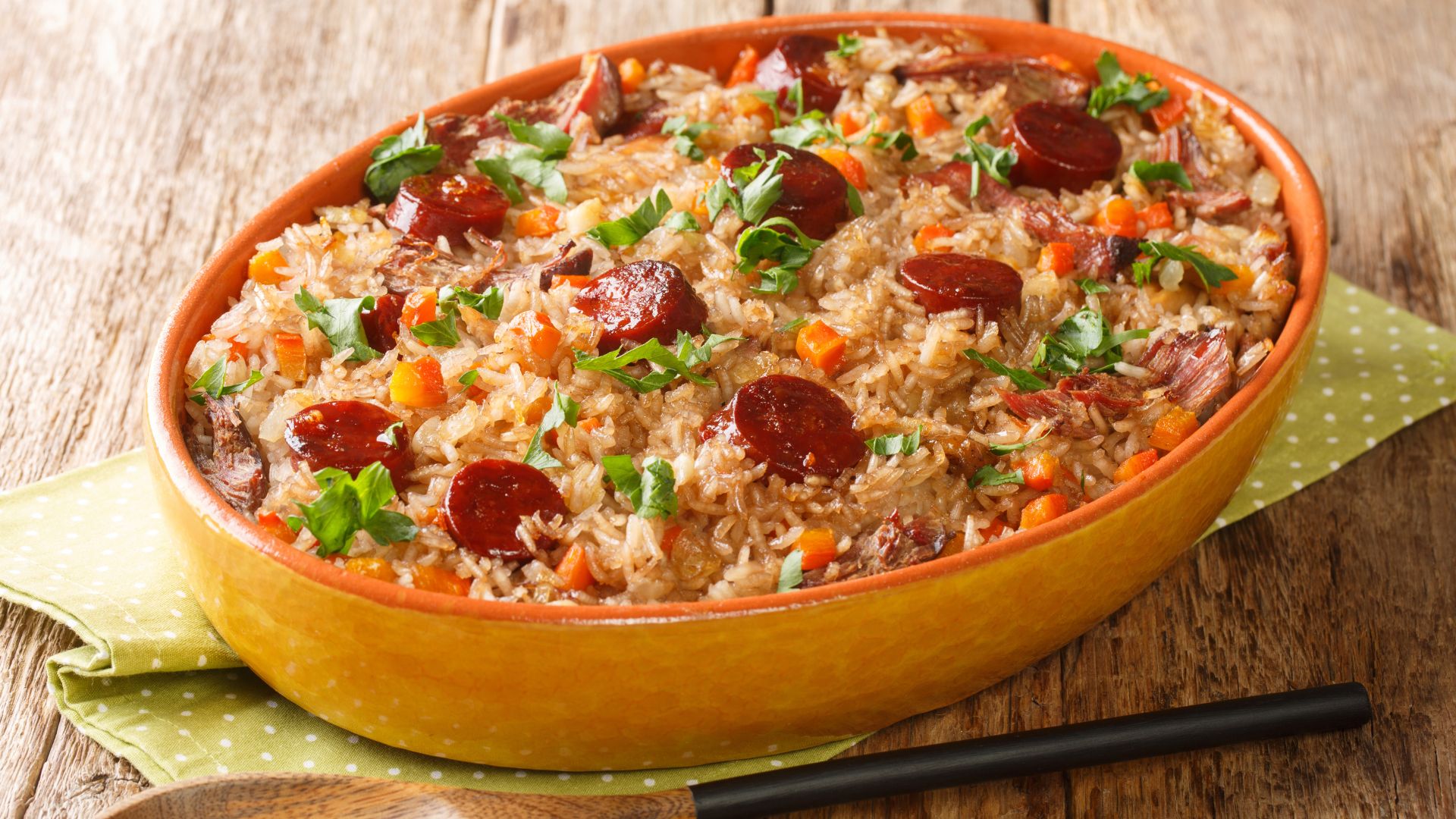 A close-up, overhead shot of Arroz de Pato in an oval baking dish, showing rice mixed with duck meat, chorizo slices, carrots, and fresh parsley. The dish is presented on a wooden table with a polka-dotted green napkin alongside a wooden spoon.