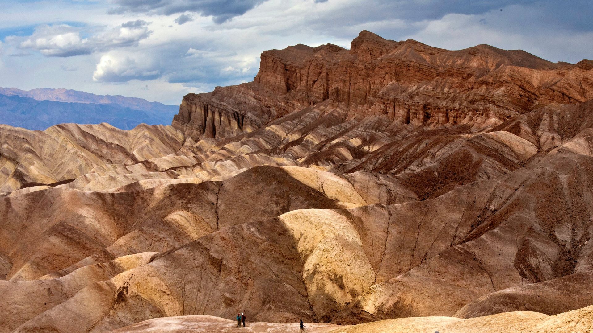 A panoramic view of the colorful, eroded badlands of Artist's Palette in Death Valley National Park, California, with a vast expanse of multi-hued hills and a distant mountain range under a partly cloudy sky, featuring a few small figures of people at the base of the hills providing a sense of scale.