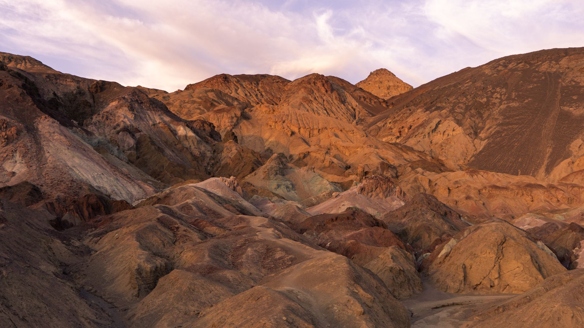 A panoramic view of the colorful, eroded badlands of Artist's Palette in Death Valley National Park, California, with varying shades of red, orange, brown, and green mineral deposits on the slopes under a partly cloudy sky.