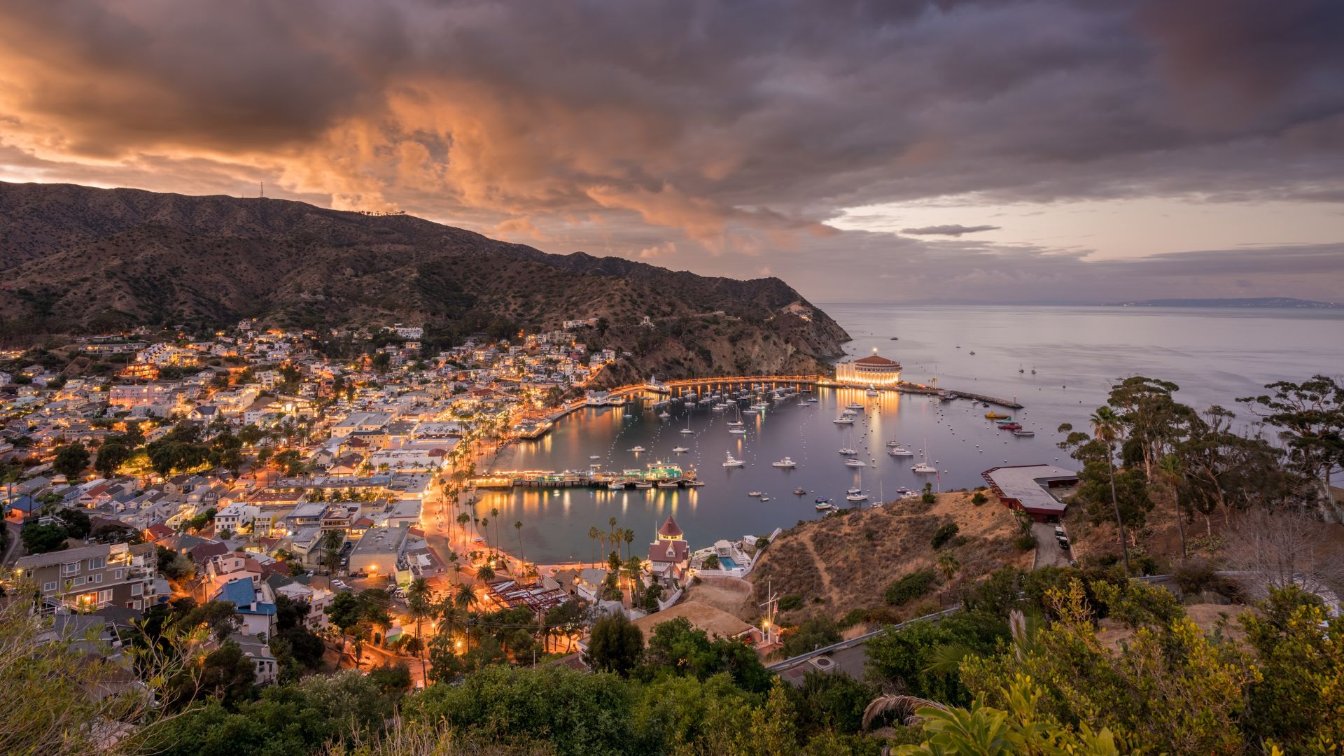 A high-angle panoramic view of Avalon Bay on Catalina Island at dusk, featuring the illuminated town nestled along the crescent-shaped harbor filled with boats, with the iconic Catalina Casino visible on the right, all set against a backdrop of rolling hills and a dramatic, cloud-filled sky with hints of orange from the sunset.