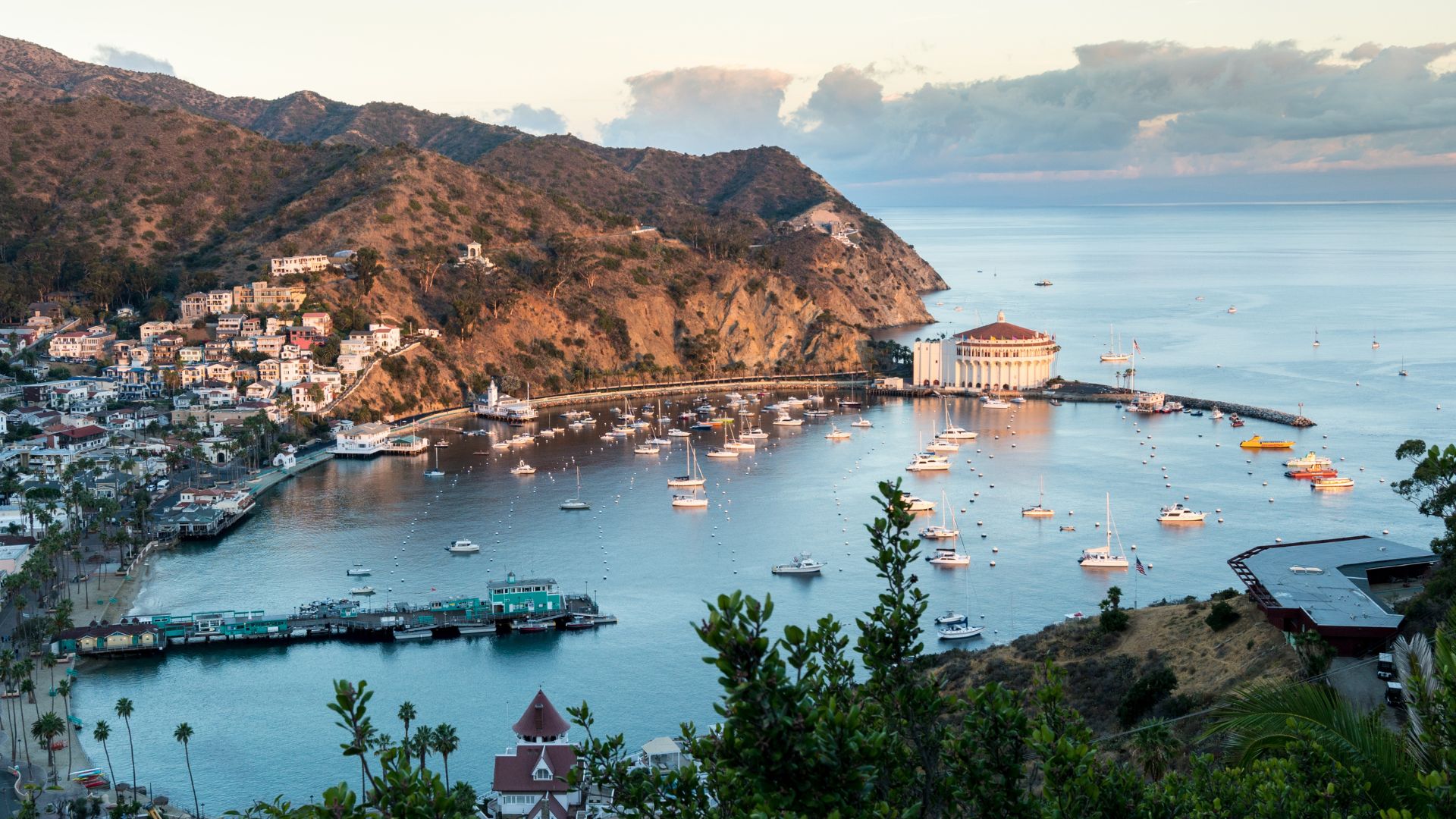 A panoramic view of Avalon Bay on Santa Catalina Island, California, at dusk, showing the harbor filled with boats, the town nestled along the hillside, and the prominent circular Catalina Casino building on the right, all under a partially cloudy sky.