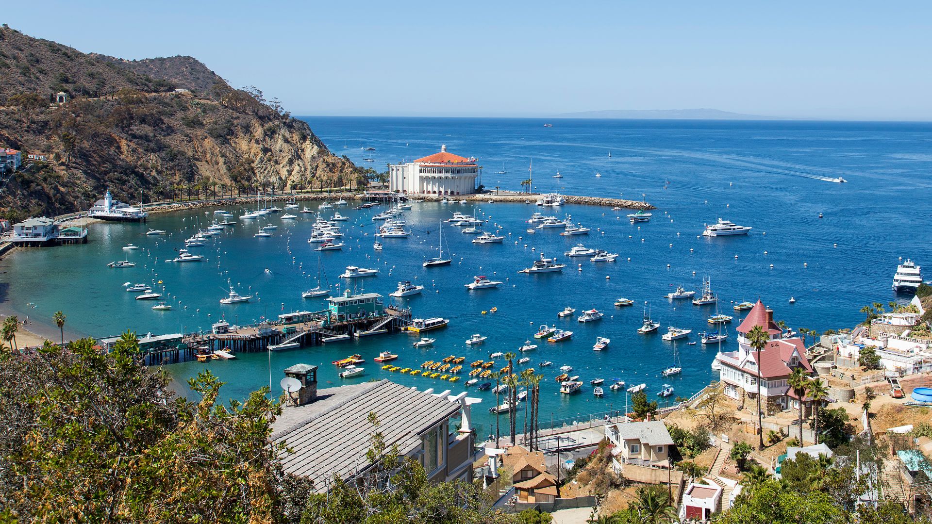 A panoramic view of Avalon Bay on Santa Catalina Island, California, featuring the iconic circular Catalina Casino building at the center, surrounded by numerous boats and yachts anchored in the clear blue water. The bay is framed by rugged, green-brown hillsides, with a long pier extending into the water in the foreground.