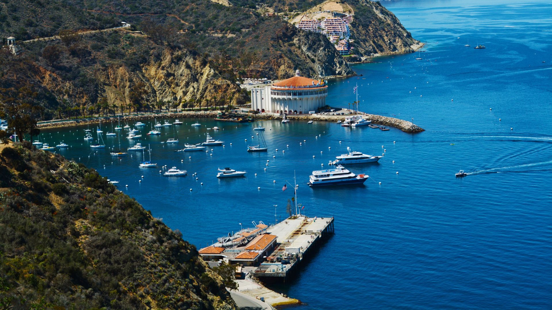 A panoramic view of Avalon Bay on Santa Catalina Island, California, at dusk, showing the harbor filled with boats, the town nestled along the hillside, and the prominent circular Catalina Casino building on the right, all under a partially cloudy sky.
