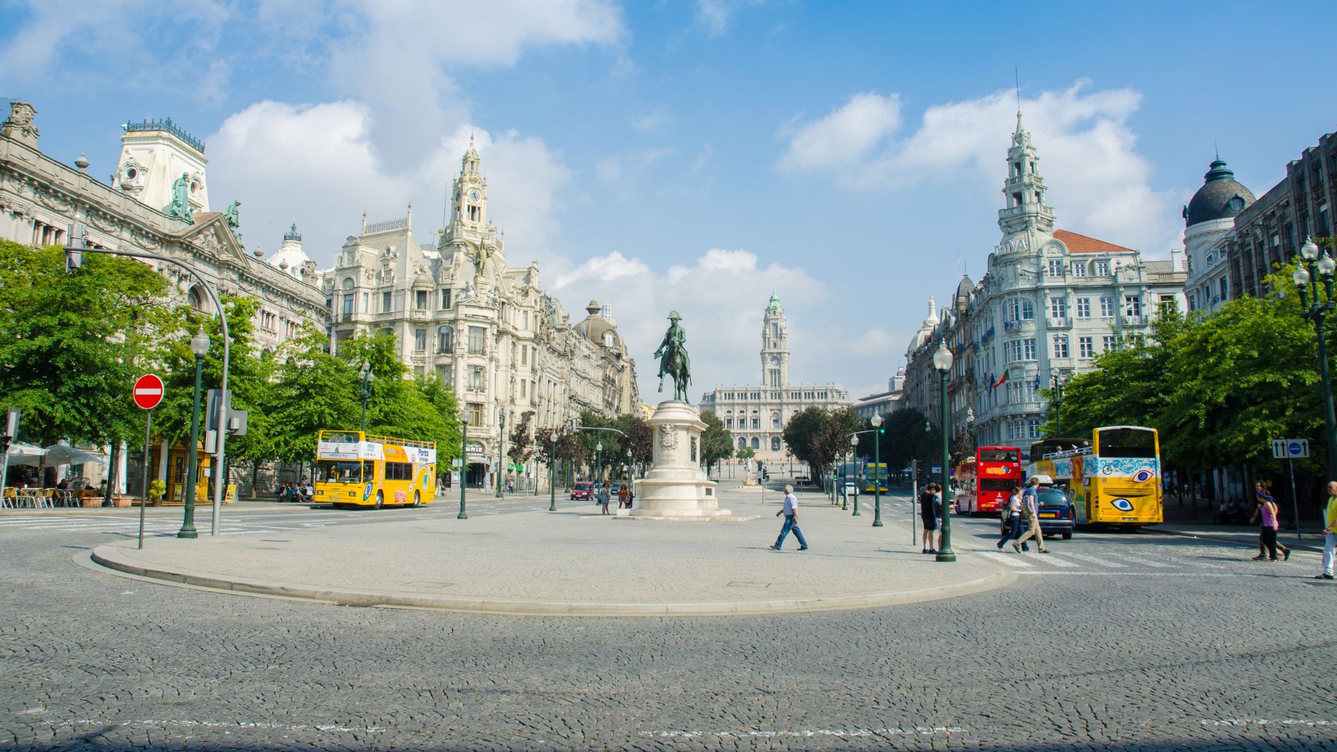 A wide, sunny view of Avenida dos Aliados in Porto, Portugal, showcasing ornate buildings, a central statue, and a bustling square.