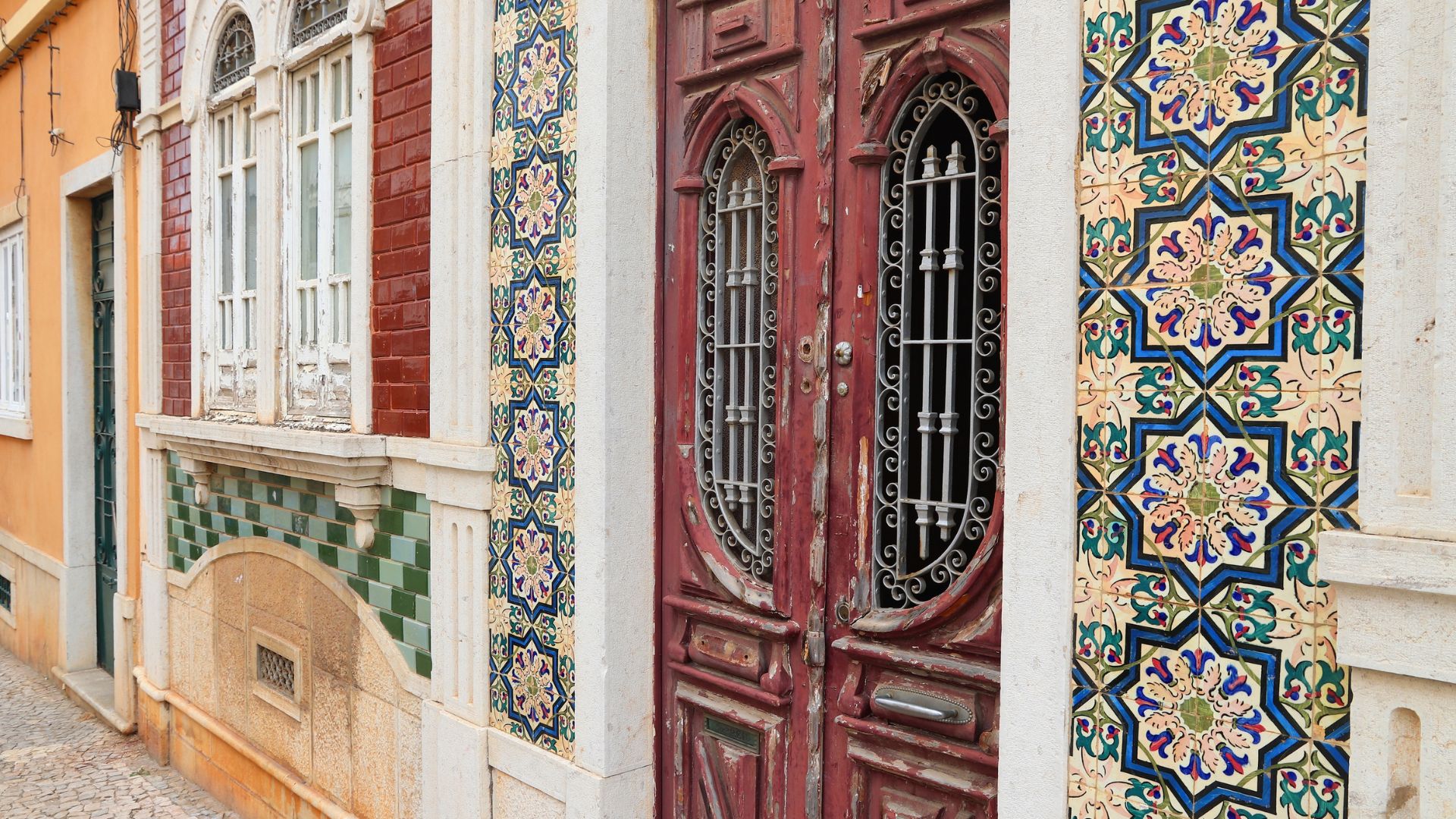 A close-up view of a building facade in Faro, Portugal, featuring traditional Portuguese red wooden doors with ornate metal grilles and decorative blue, green, and white Azulejo tiles on the walls surrounding the entrance and beneath the windows. 