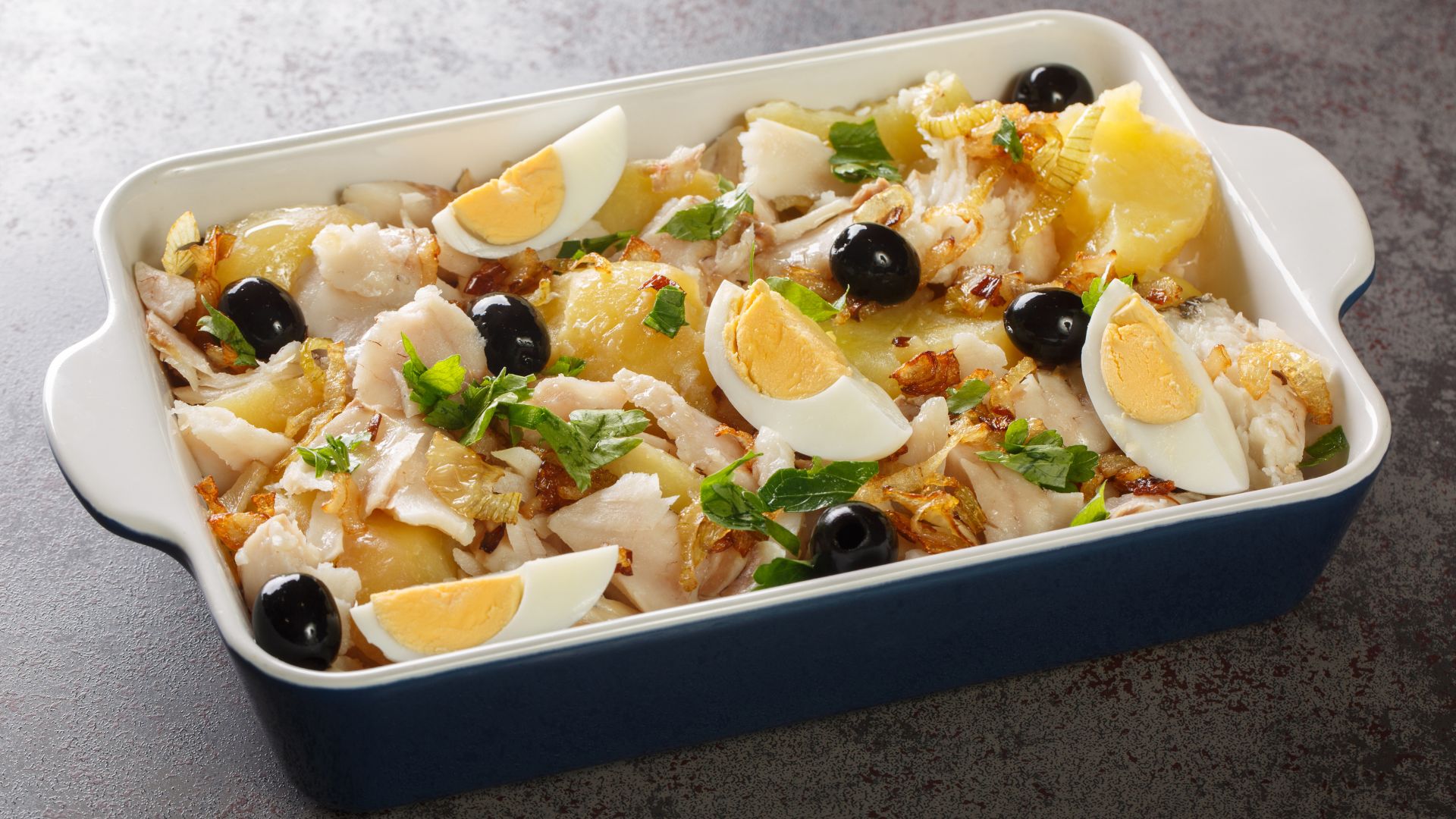 A baking dish filled with Bacalhau à Gomes de Sá, a traditional Portuguese casserole featuring flaked salted cod, sliced potatoes, onions, black olives, and garnished with quartered hard-boiled eggs and fresh parsley. 