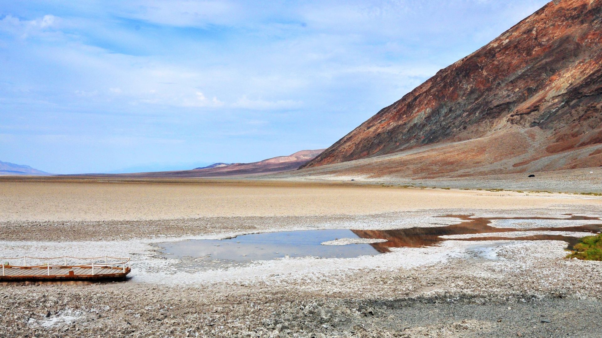 A wide, low-angle view of Badwater Basin in Death Valley National Park, featuring the vast, salt-covered flats and a small pool of water in the foreground, with a large, barren mountain slope rising sharply on the right under a blue sky with scattered clouds.