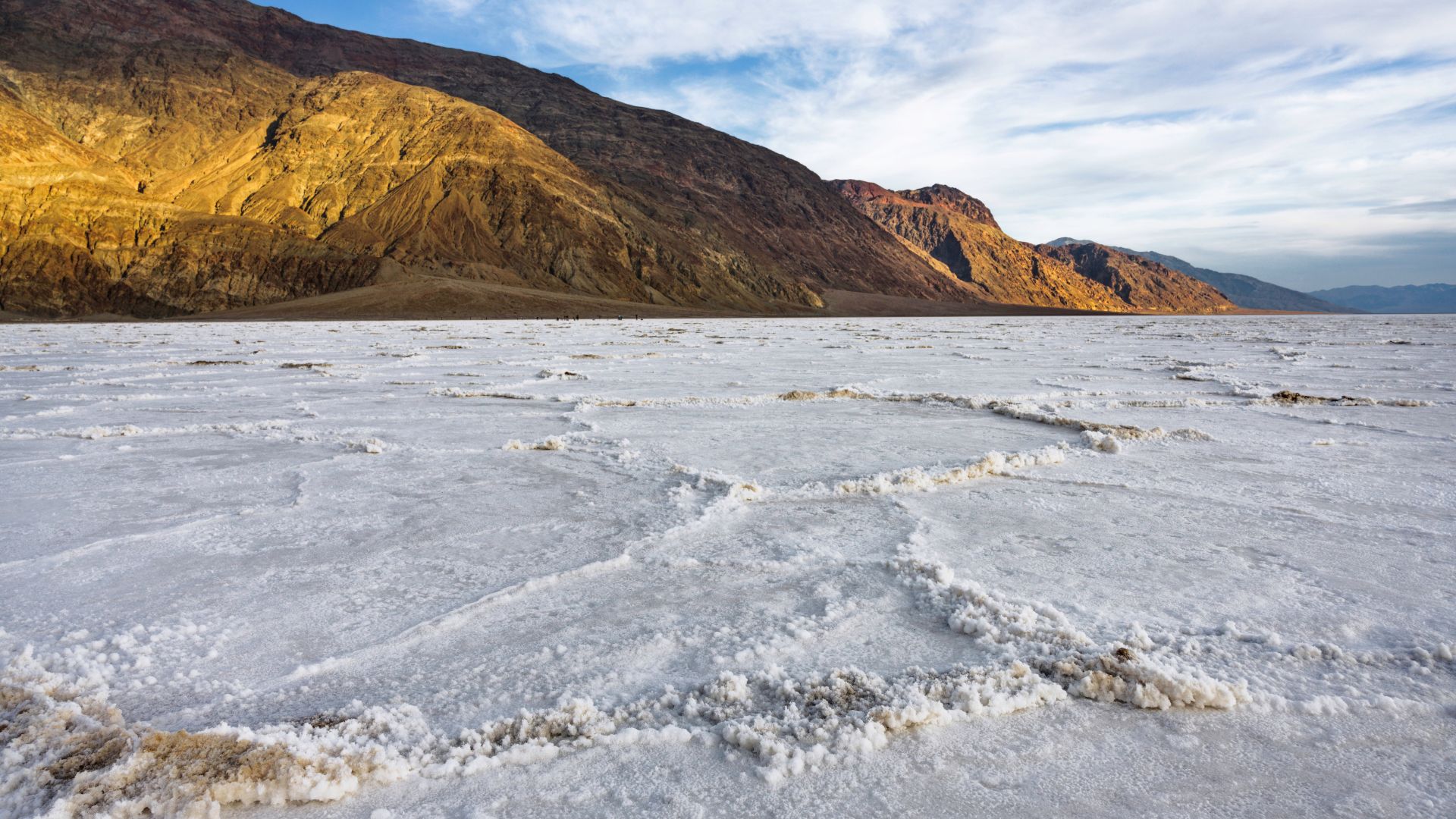 A wide-angle view of the vast, white salt flats of Badwater Basin in Death Valley National Park, California, with distinct polygonal salt formations on the ground and sunlit, rugged mountains rising in the background under a partly cloudy sky.
