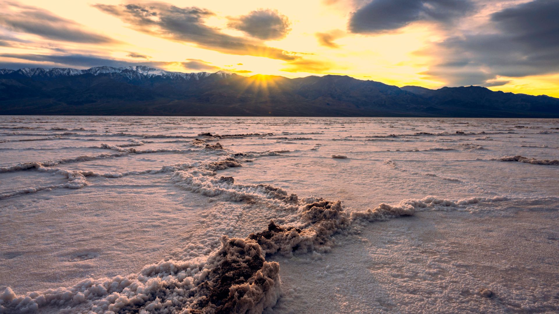 Sunset over the vast salt flats of Badwater Basin in Death Valley National Park, California, with distant mountains silhouetted against a vibrant sky.