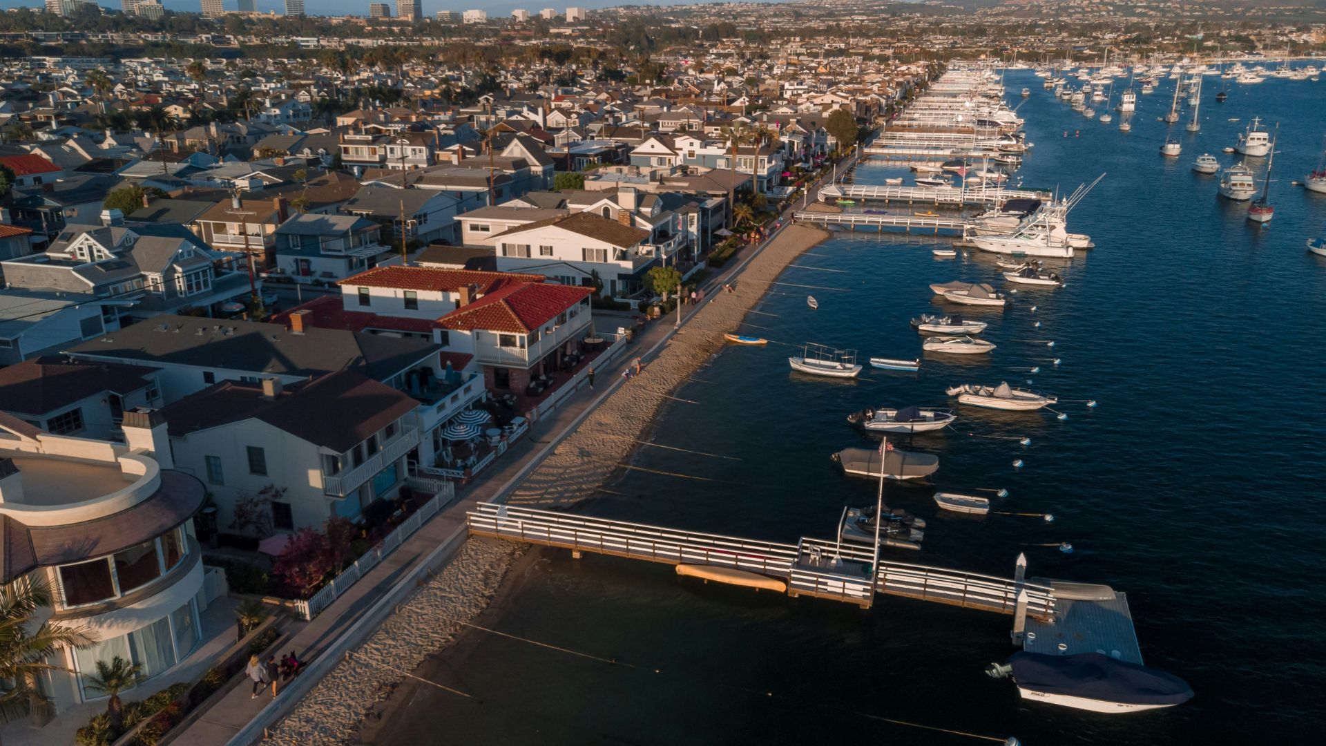 Aerial view of Balboa Island in Newport Beach, California, showcasing waterfront homes, sandy beaches, and numerous boats docked in the scenic harbor under a clear sky.