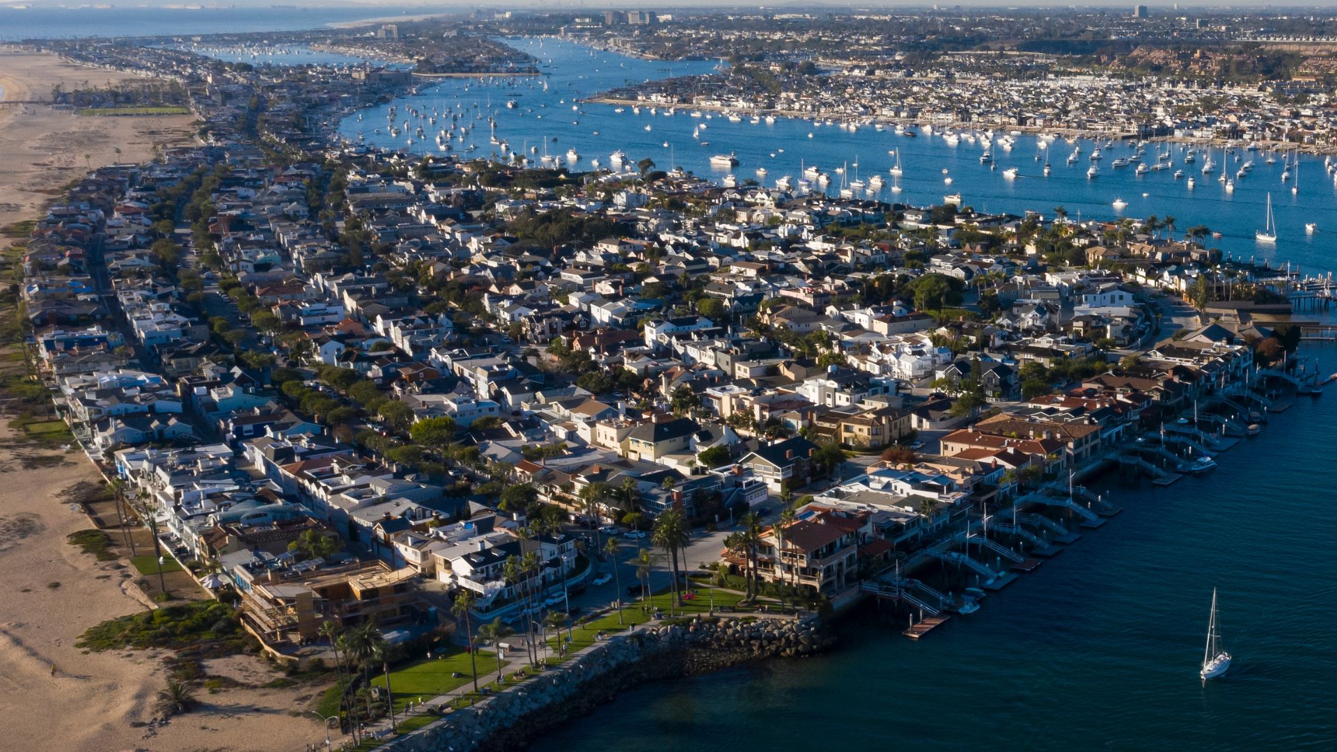 Aerial view of Balboa Island in Newport Beach, California, showcasing waterfront homes, a bustling harbor filled with boats, and the surrounding coastline.