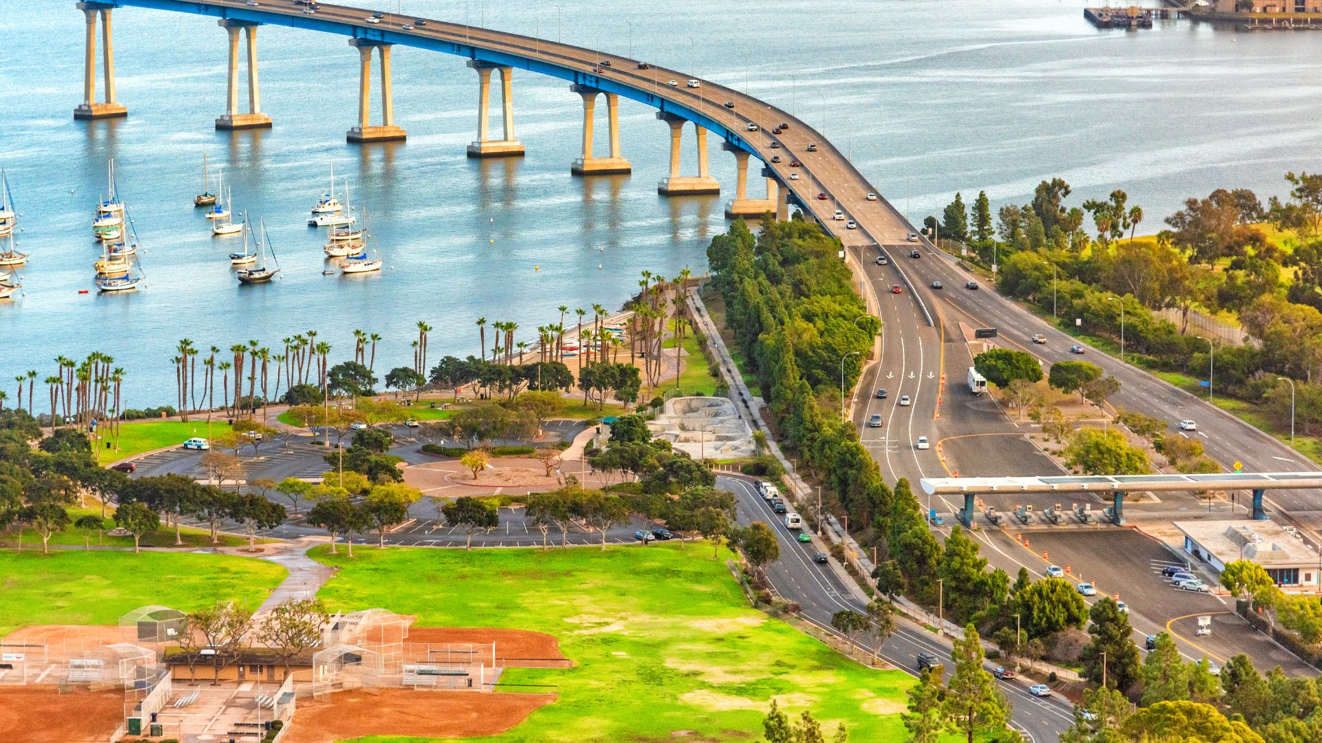  An aerial view of the San Diego–Coronado Bridge spanning across the San Diego Bay, with sailboats on the water and a park and roads visible on the land in the foreground.