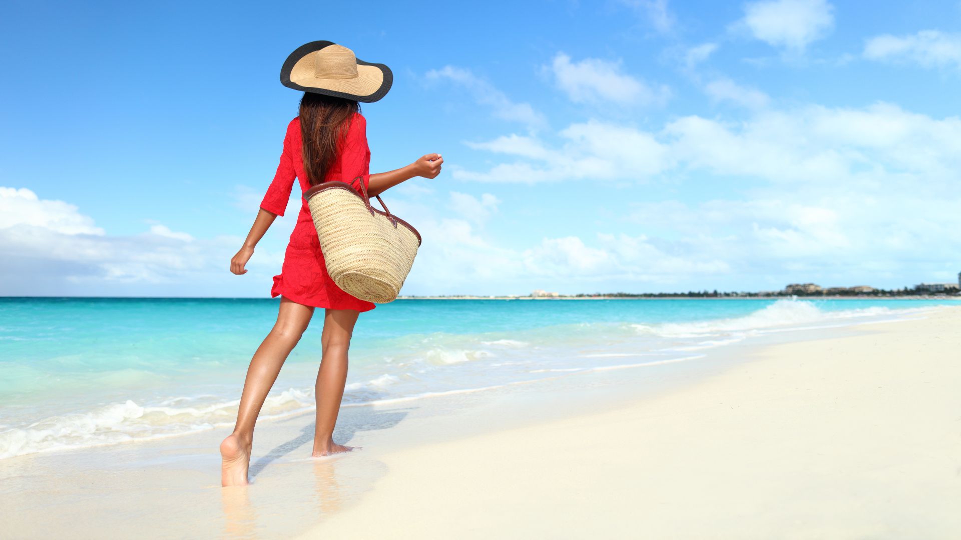 Woman wearing red dress with a bag and hat at the beach.
