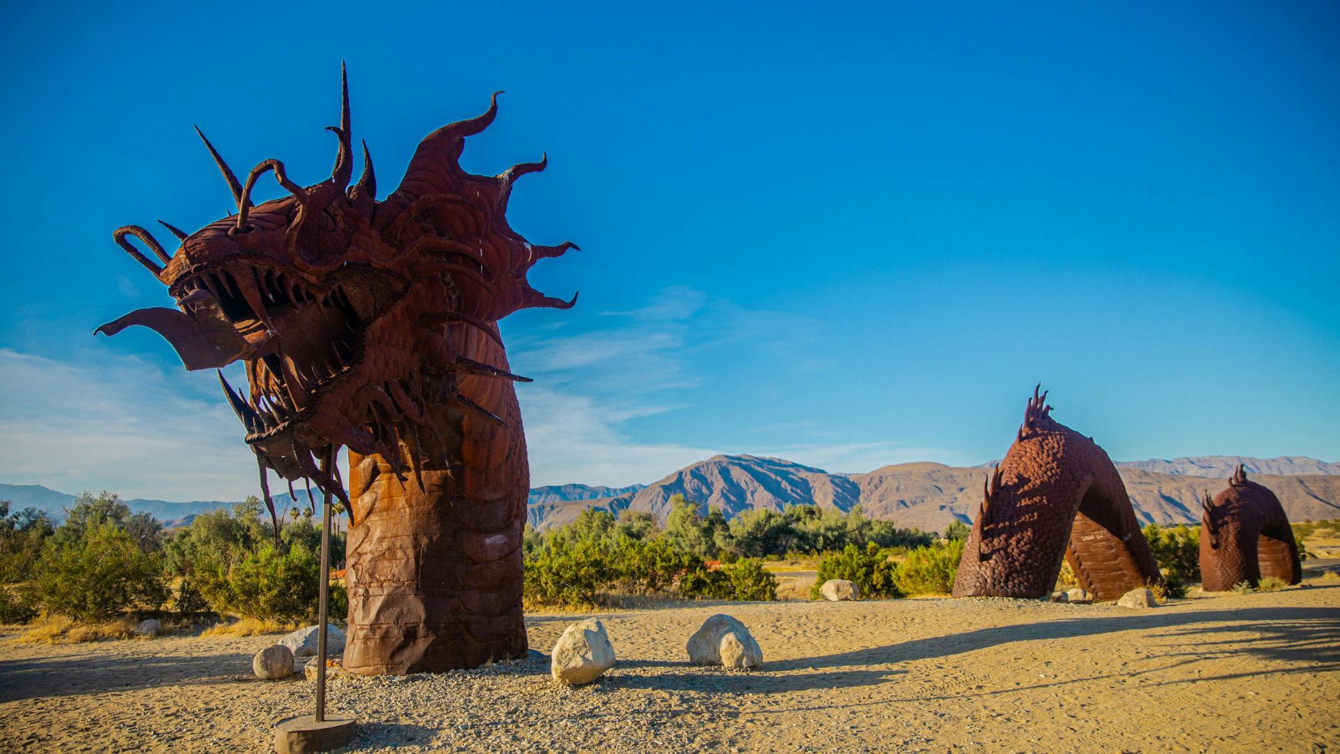 A wide shot of a desert landscape under a clear blue sky, featuring large, weathered metal sculptures. In the foreground on the left, a towering, intricate sculpture of a dragon's head and neck rises from the sandy ground. To the right, two smaller, rounded, organic-shaped metal structures are visible in the distance, resembling coiled serpents or abstract forms. The background reveals a range of arid mountains under the bright sky.