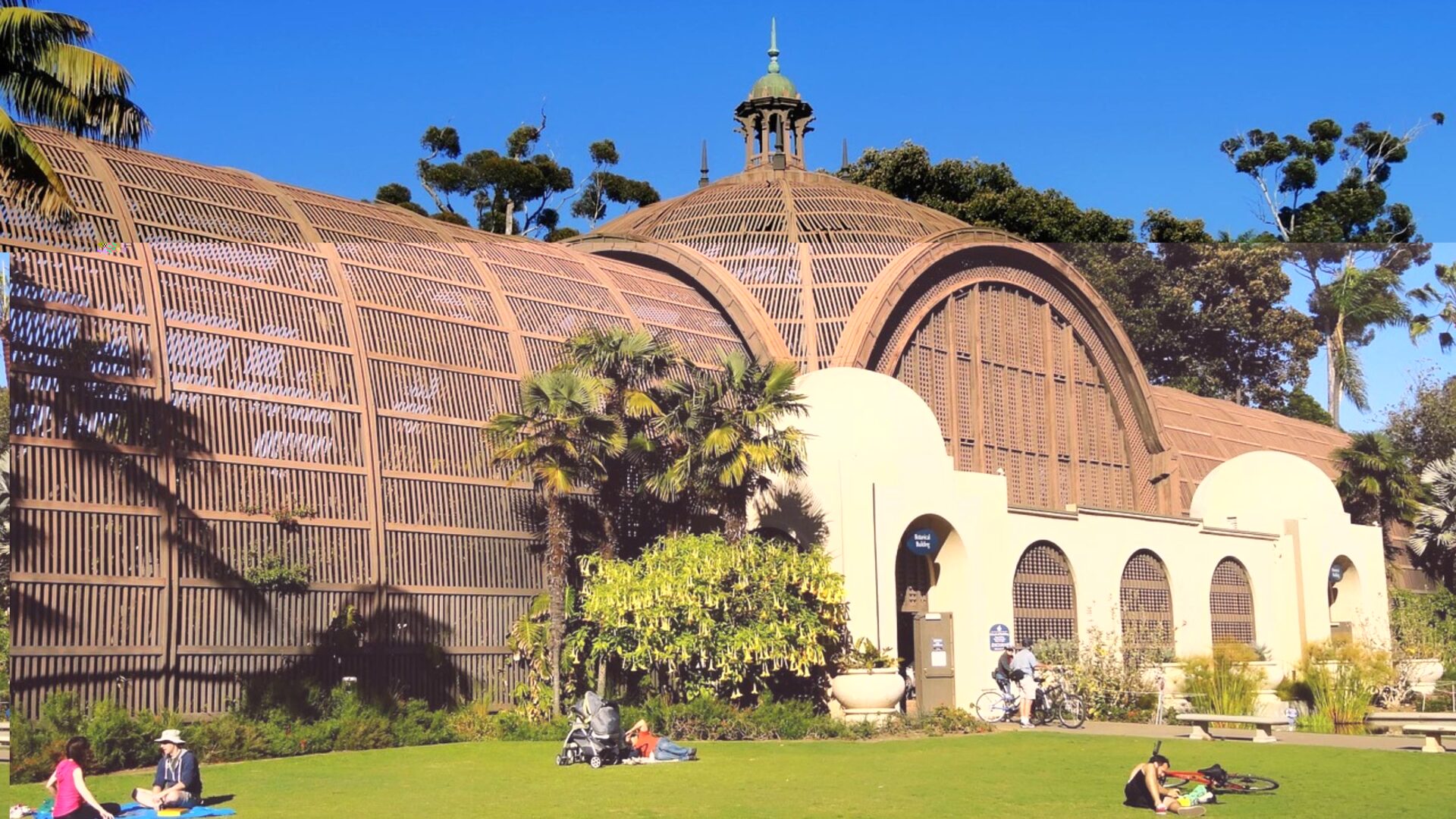 A wide shot of the iconic Botanical Building in Balboa Park, San Diego, with its distinctive arched wooden lath structure and surrounding green lawn where people are relaxing.