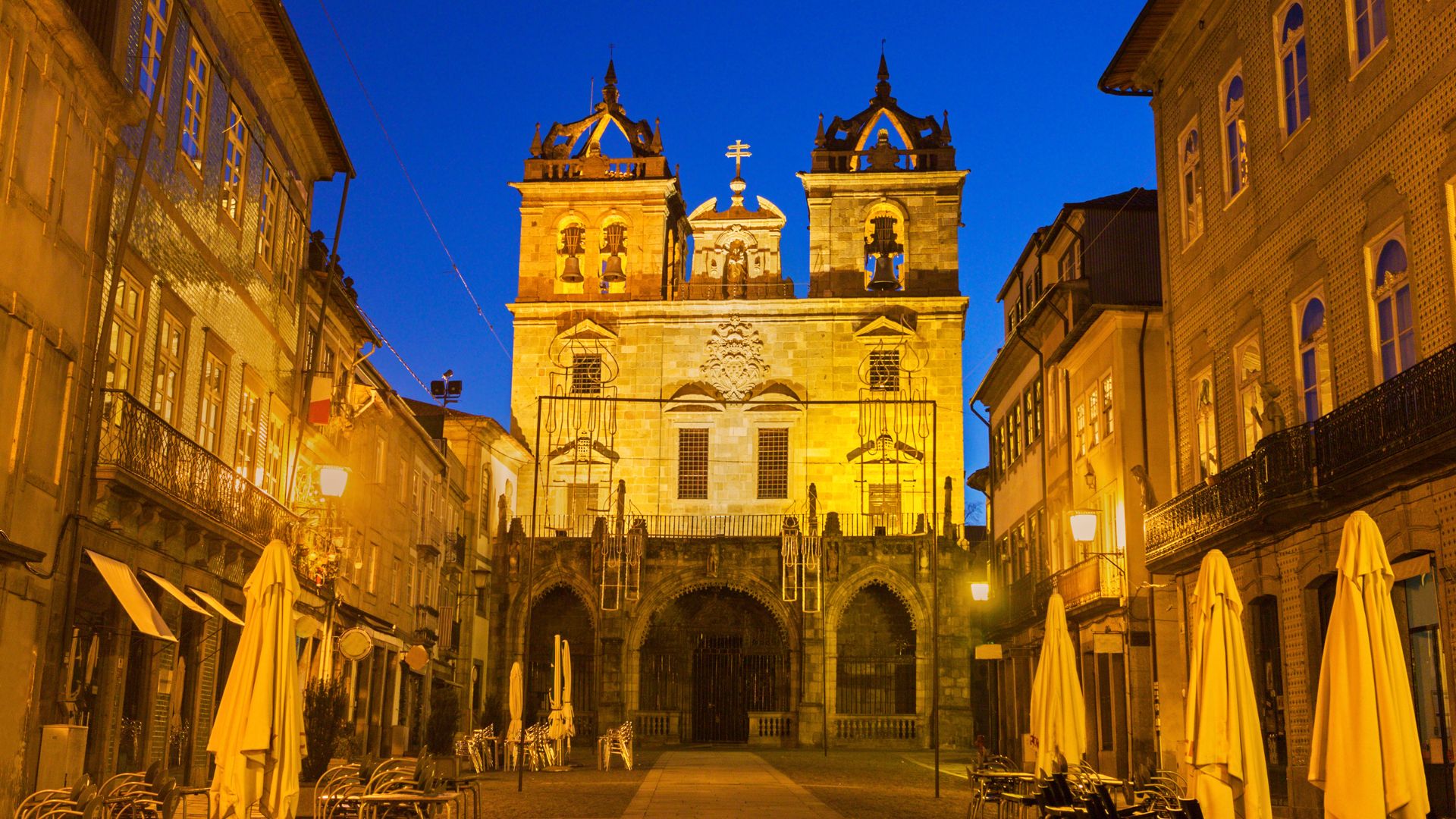 A night view of the illuminated Braga Cathedral in Portugal, with narrow streets and buildings on either side, and yellow umbrellas from outdoor cafes visible in the foreground.
