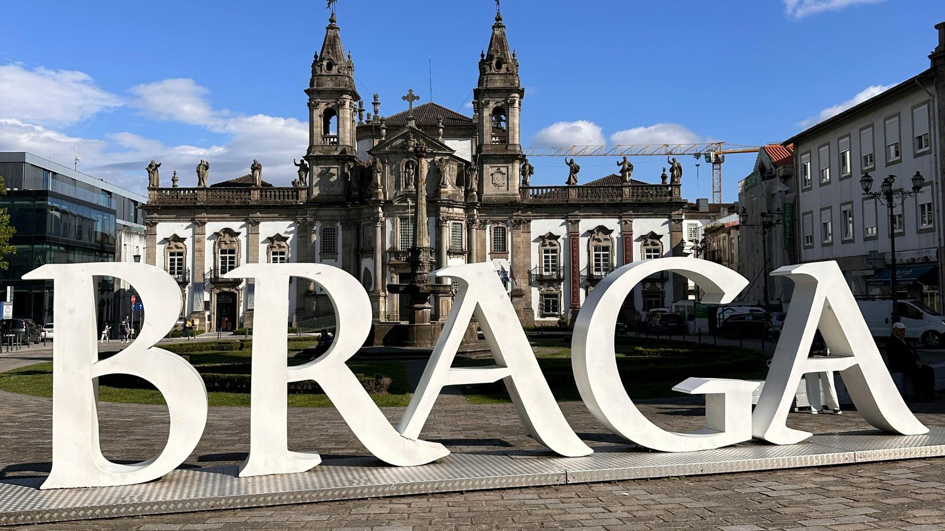 Large white letters spelling "BRAGA" in a public square in Portugal, with the historic Braga Cathedral visible in the background under a blue sky.