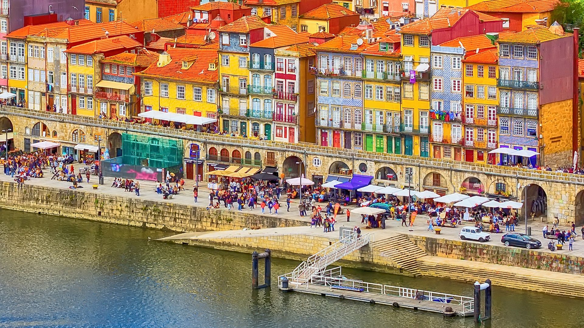 A high-angle view of Cais da Ribeira in Porto, Portugal, showing colorful buildings along the Douro River, with people strolling along the promenade and a bridge in the background.