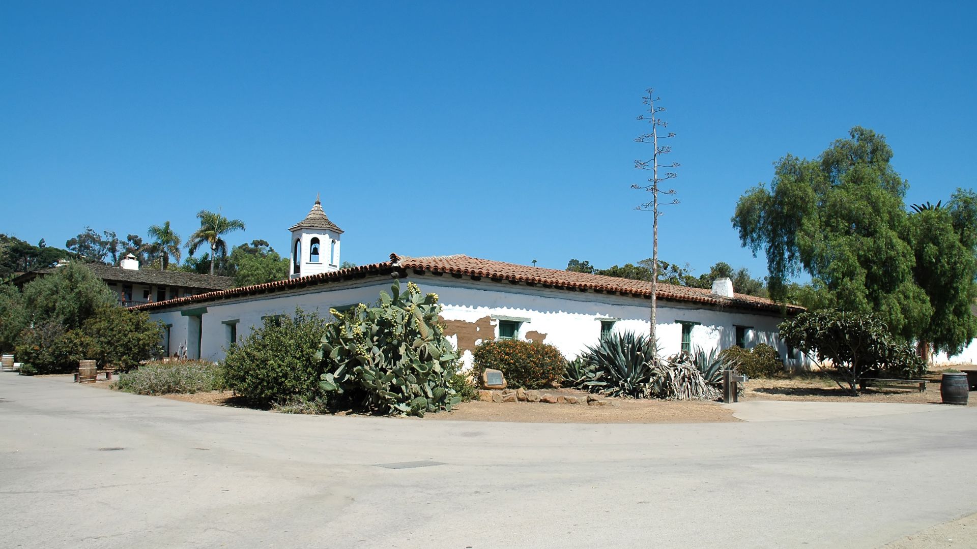A historic, single-story, white adobe building with a red tile roof and a bell tower, surrounded by lush green foliage and cacti under a clear blue sky in Old Town San Diego.