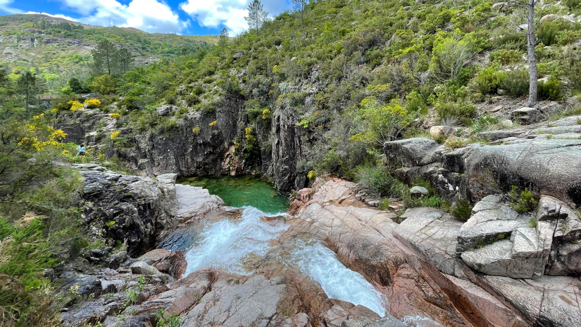 A cascading waterfall flows into a clear, green pool surrounded by lush, rocky terrain and verdant mountains under a blue sky.