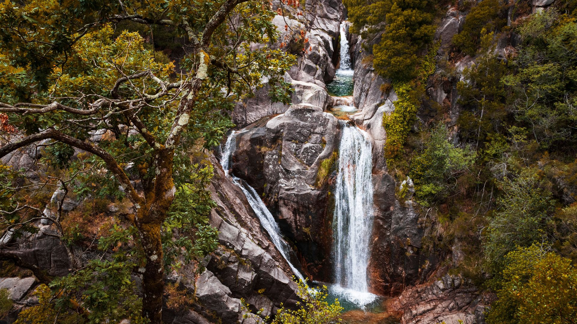 A multi-tiered waterfall cascades down rocky cliffs surrounded by lush green and autumnal foliage in a forested area.