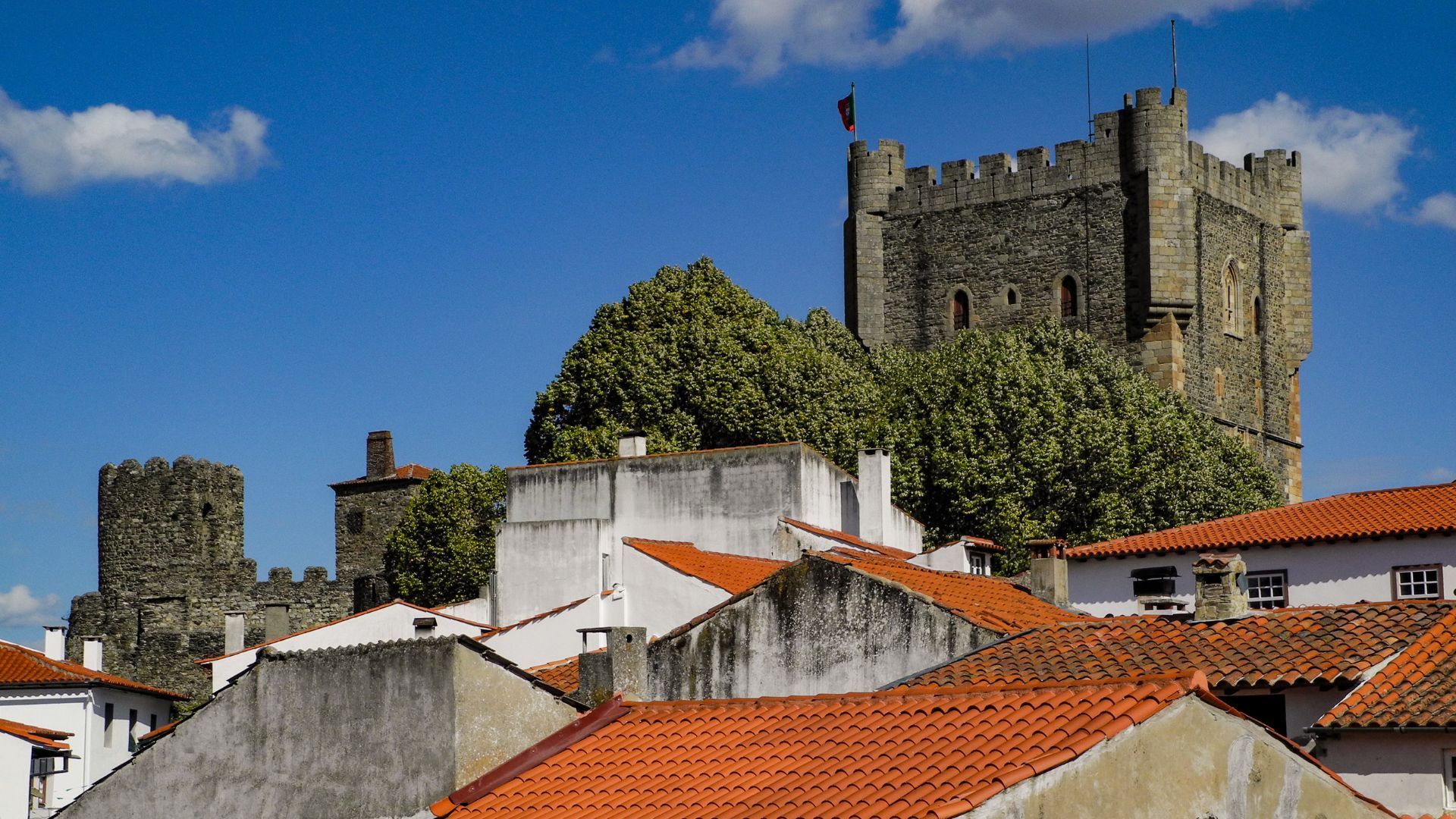 A medieval stone castle with tall towers and battlements stands prominently on a hill, overlooking a cluster of buildings with red-tiled roofs under a clear blue sky.