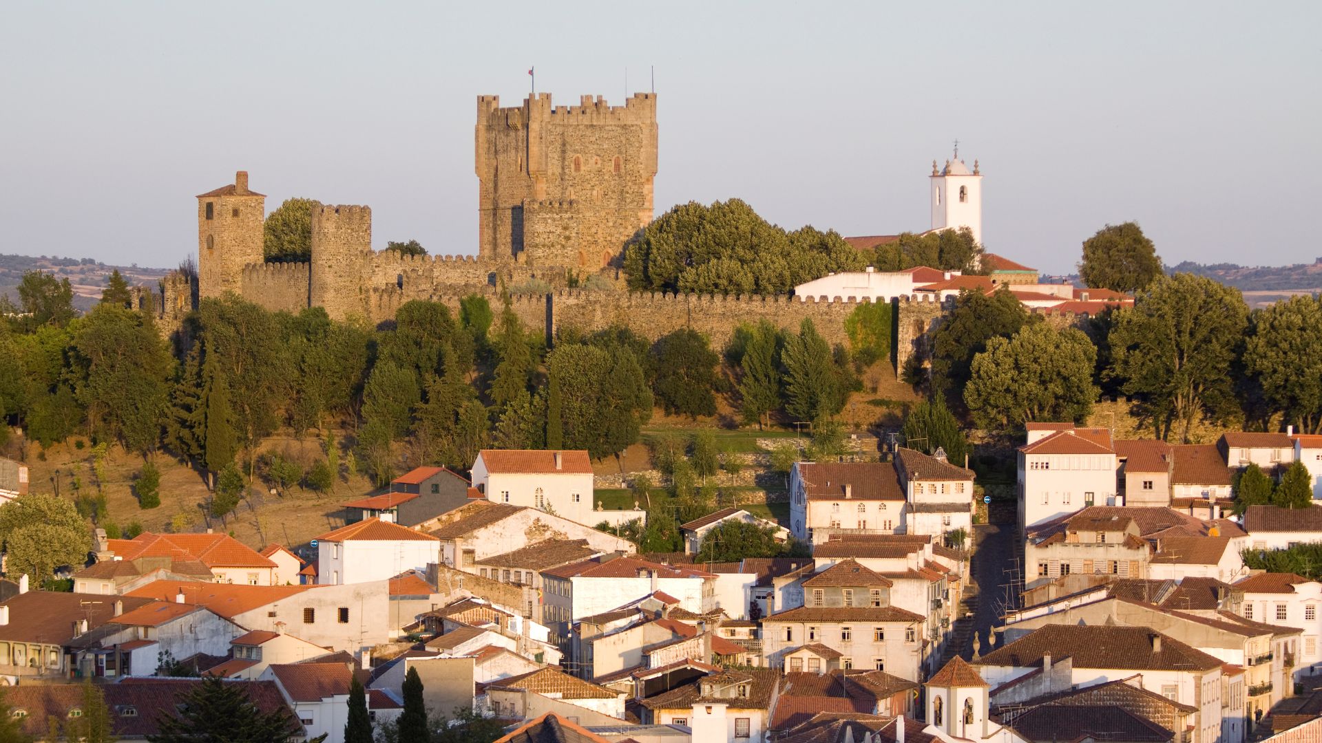 A medieval castle with stone walls and towers sits atop a hill, overlooking a town with red-tiled roofs nestled in a valley, surrounded by green trees under a clear sky.