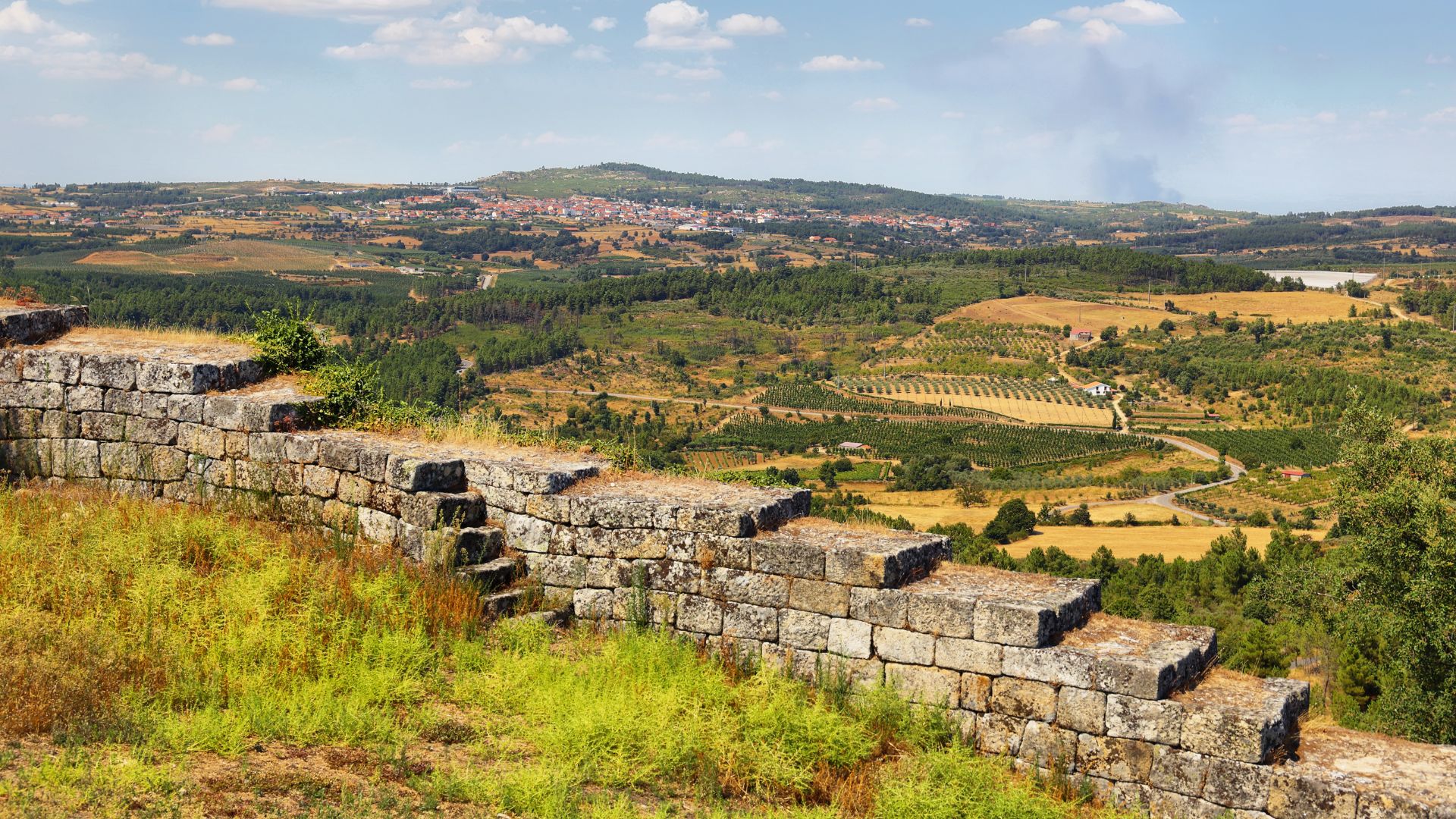A panoramic view showcasing the ancient stone walls of the Castle of Carrazeda de Ansiães in the foreground, overlooking a vast, rolling landscape of fields, vineyards, and a distant village under a partly cloudy sky in Portugal.