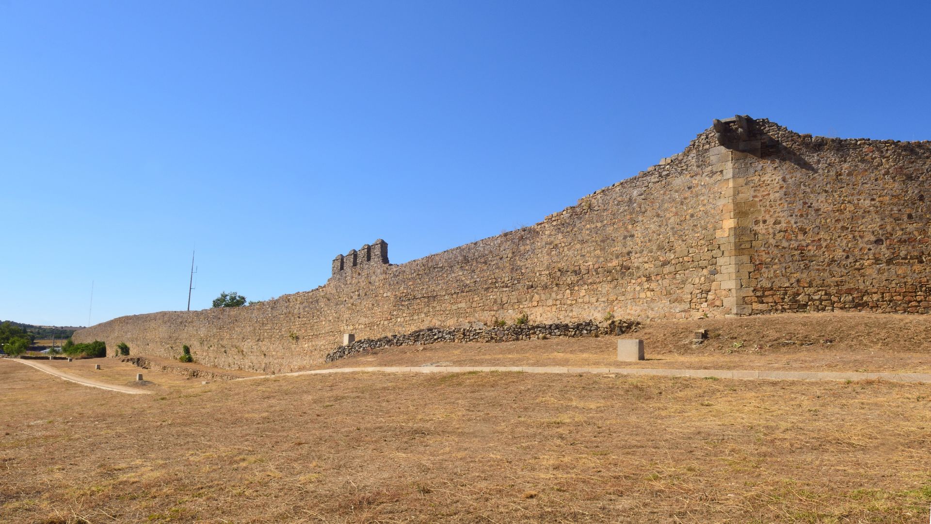 A long, ancient stone wall under a clear blue sky, extending across a dry, grassy landscape.