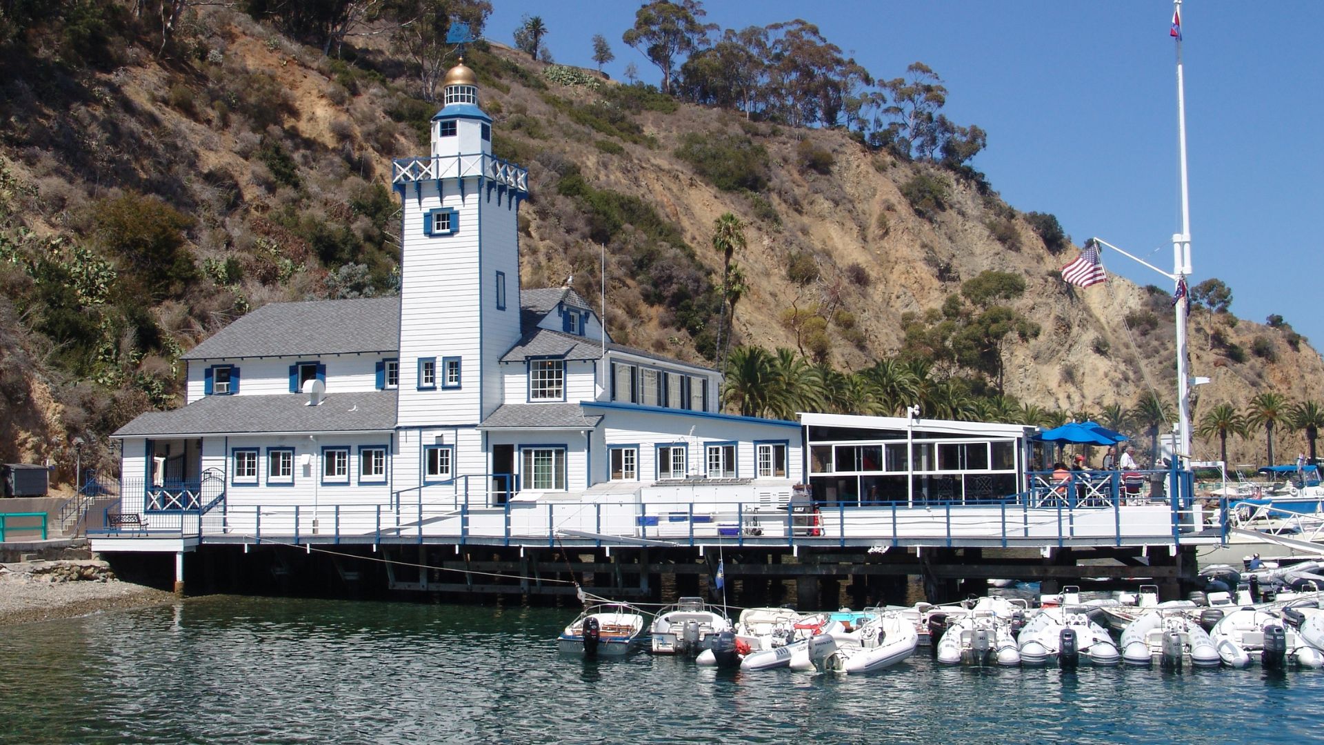 A white and blue multi-story building with a prominent tower, identified as the Catalina Island Yacht Club, situated on a pier over calm water with numerous boats docked alongside, all set against a steep, green hillside under a clear blue sky.