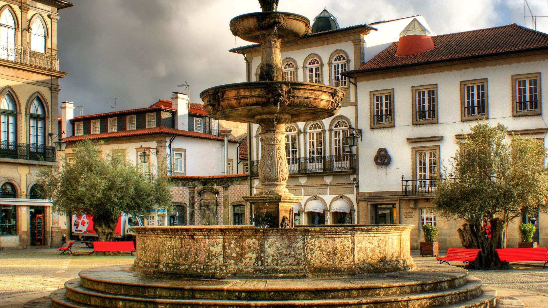 A historic stone fountain with multiple tiers in the center of a cobblestone square, surrounded by traditional white buildings with red-tiled roofs and green trees, under a cloudy sky in Ponte de Lima, Portugal.