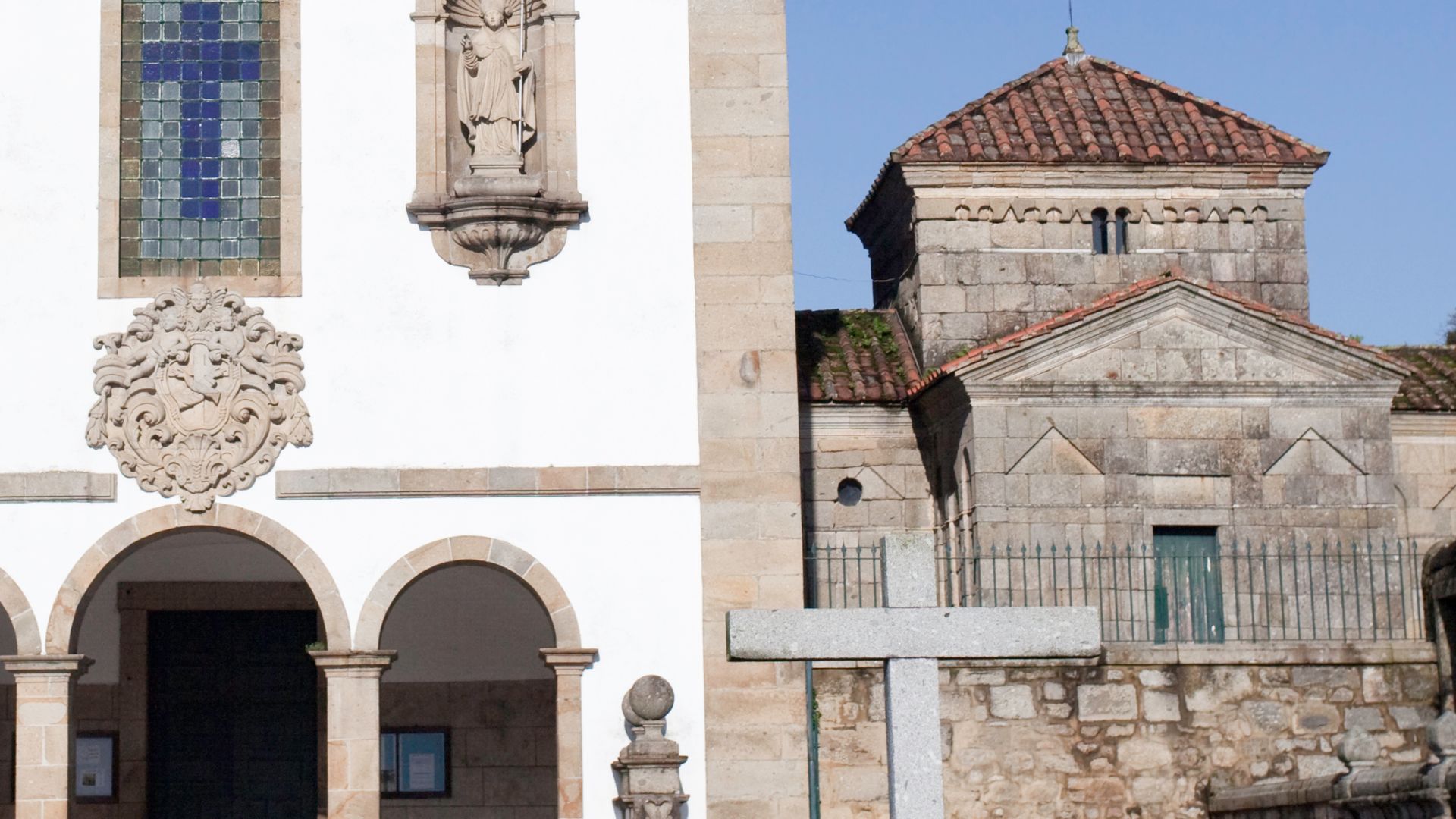 A stone chapel with a tiled roof and a cross in the foreground, set against a clear blue sky.