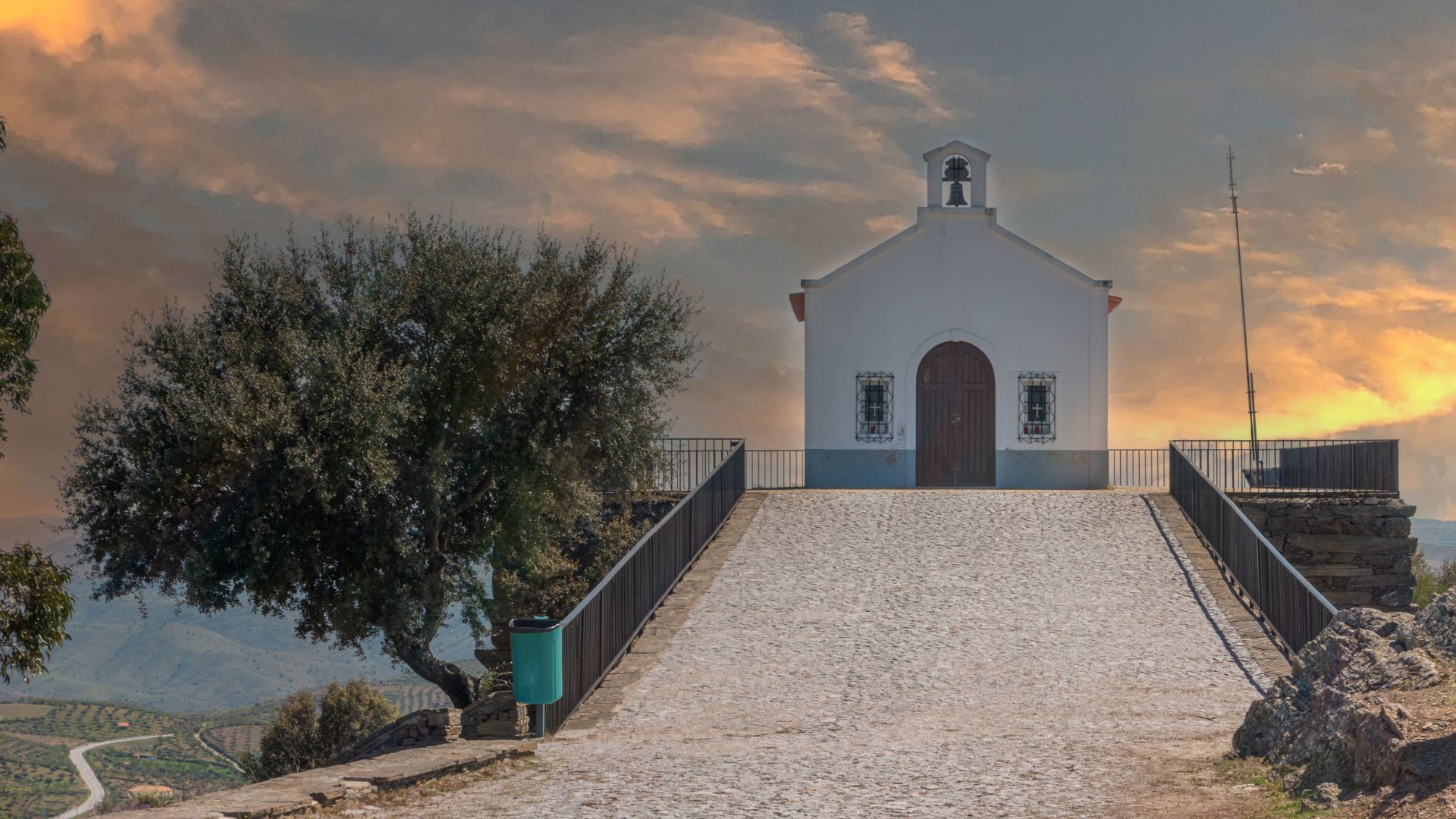 A white chapel with a bell tower at the top of a stone path, flanked by an olive tree on the left and a stone cross on the right, under a dramatic sunset sky in Castelo Melhor, Portugal.