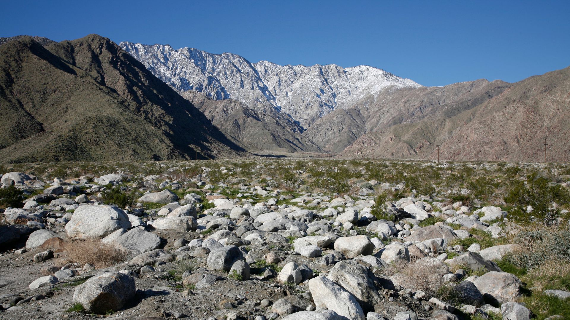 A wide shot of a rocky, desert landscape with large boulders in the foreground and a prominent mountain range with snow-capped peaks in the background under a clear blue sky, depicting Chino Canyon in the San Jacinto Mountains near Palm Springs, California.