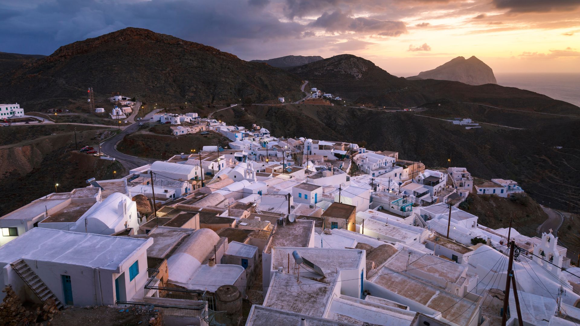 Anafi, Greece: Picturesque Chora village with white-washed houses on a hillside overlooking the Aegean Sea at sunset.
