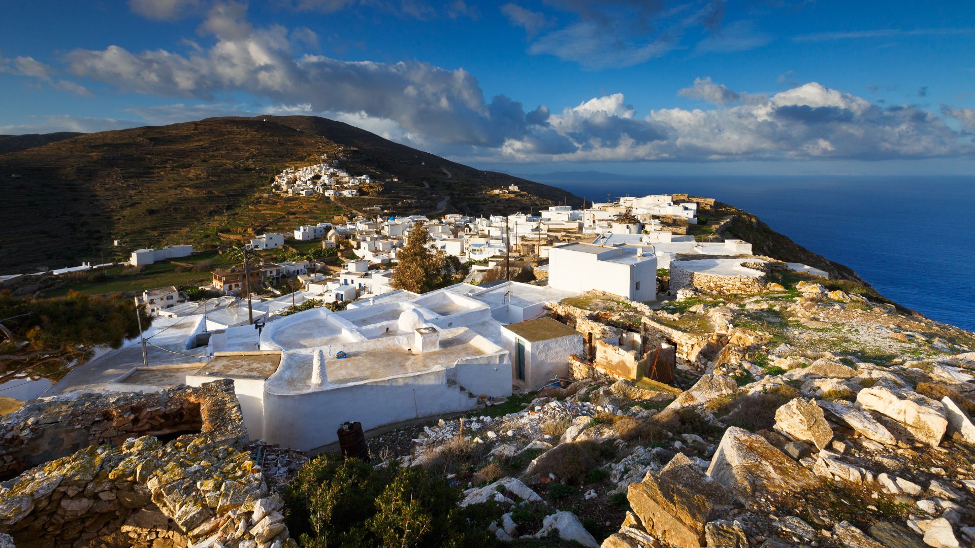 Chora village on Sikinos island, Greece, featuring traditional white Cycladic architecture against a mountainous backdrop and the Aegean Sea.