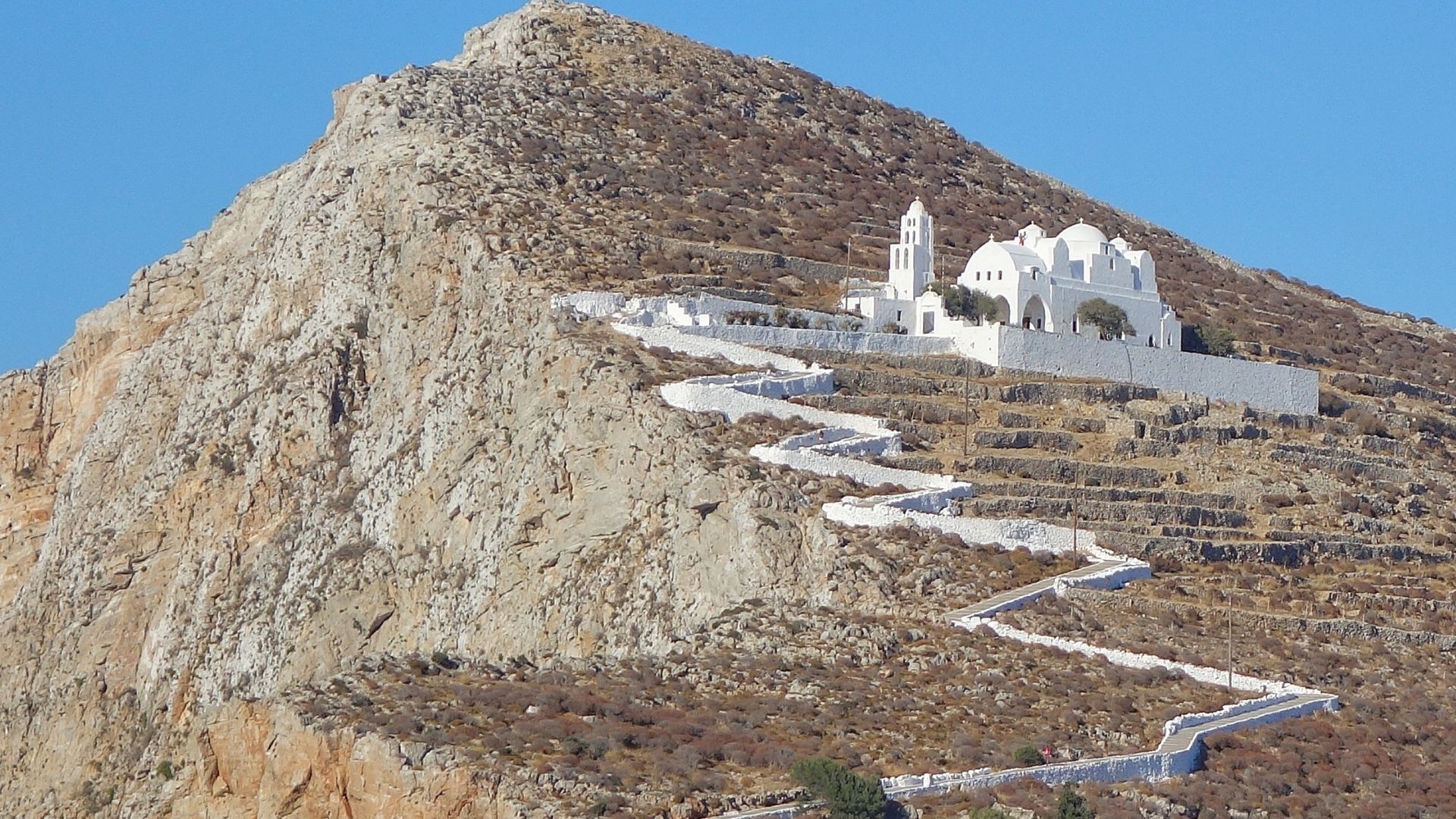 Whitewashed church on a hill with a winding path leading up to it in Folegandros, Greece.